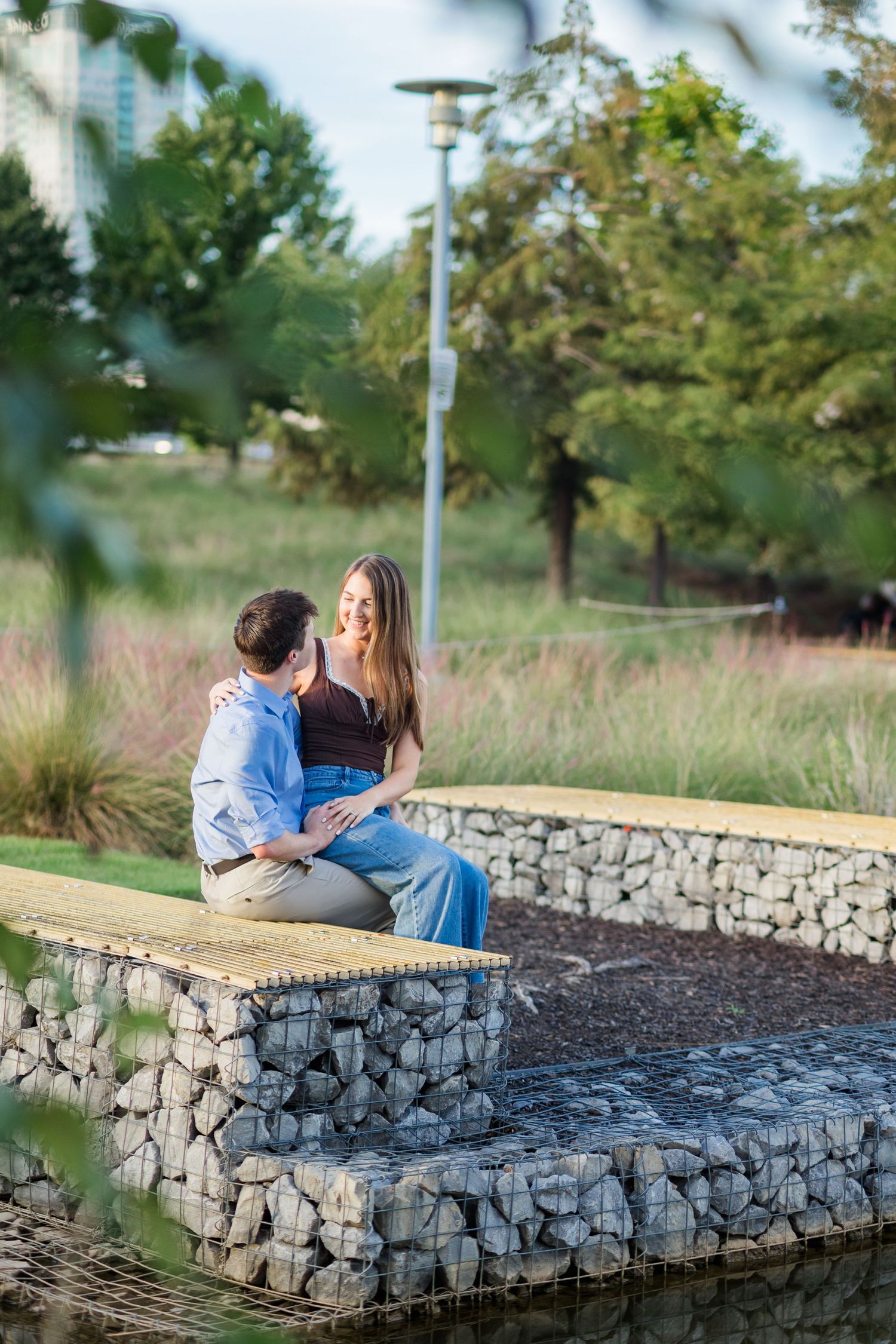 Romantic couple sitting together on stone gabion wall in urban park surrounded by lush greenery.