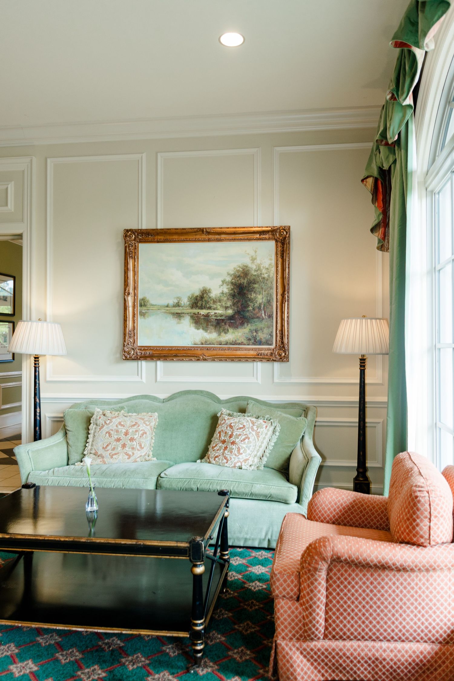 Classic living room with mint green sofa, pink armchairs, gold framed landscape painting, and white paneled walls.