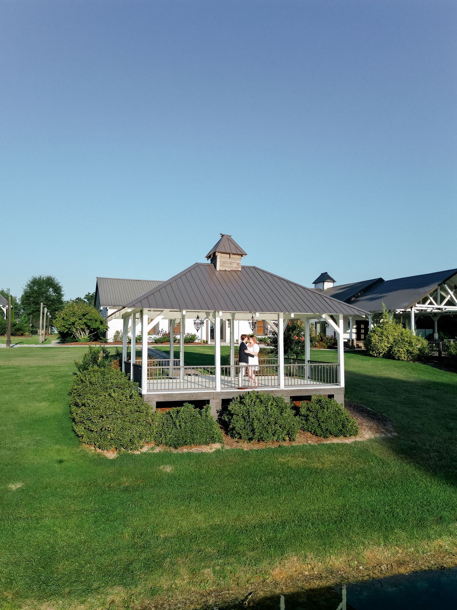 Couple embracing in a white gazebo surrounded by lush greenery at a scenic outdoor venue.