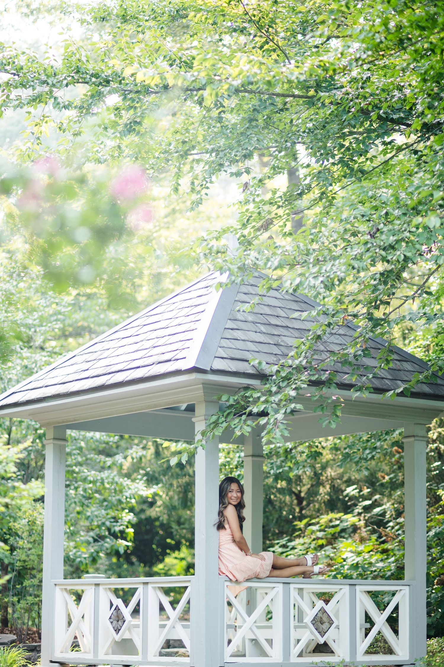 Young woman in peach dress sitting in white gazebo surrounded by lush green trees.