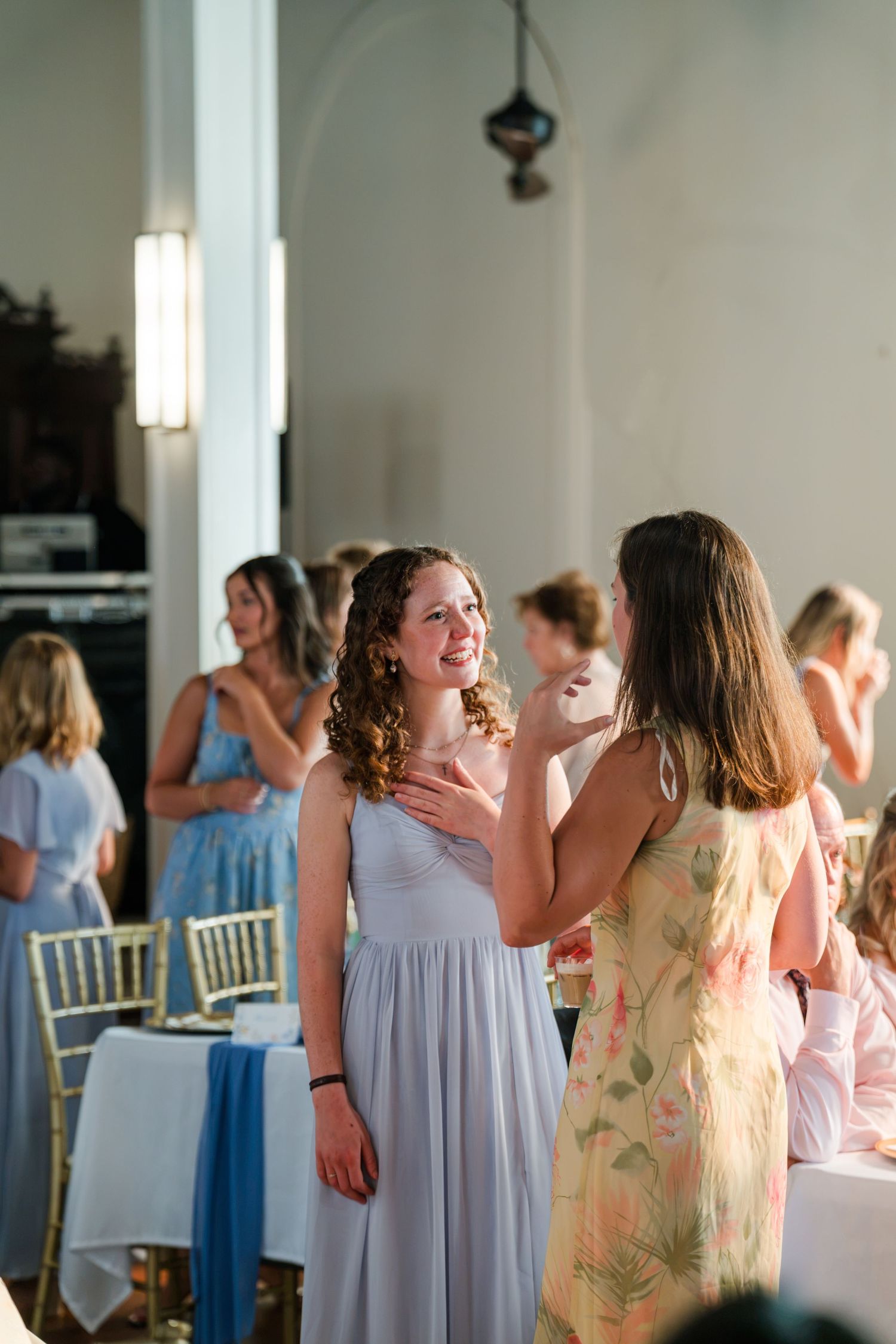 Bridesmaid in a lavender gown smiling and chatting with a guest during a sunny wedding reception indoors.