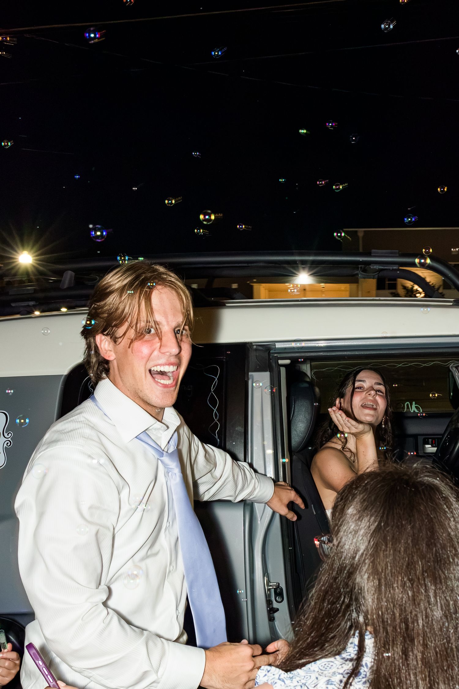 Laughing groom in white shirt and blue tie exits vehicle at night during wedding celebration.