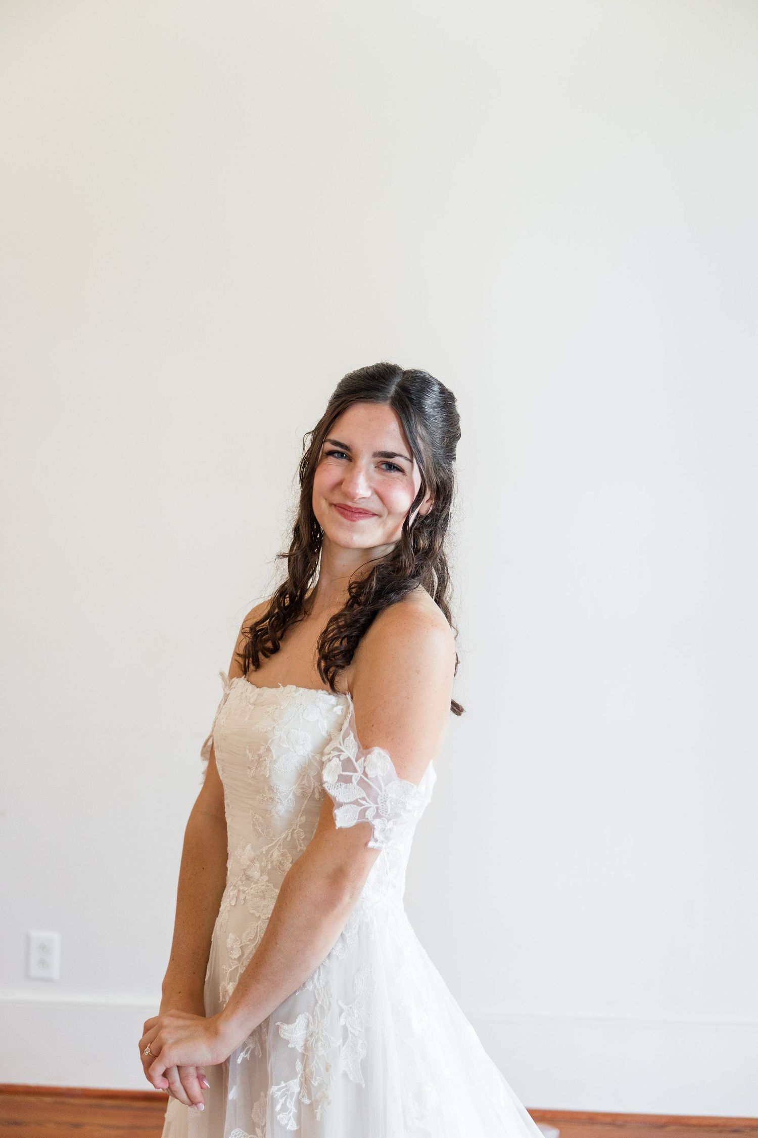 Brunette bride smiling in an off-shoulder lace wedding gown against a white wall.