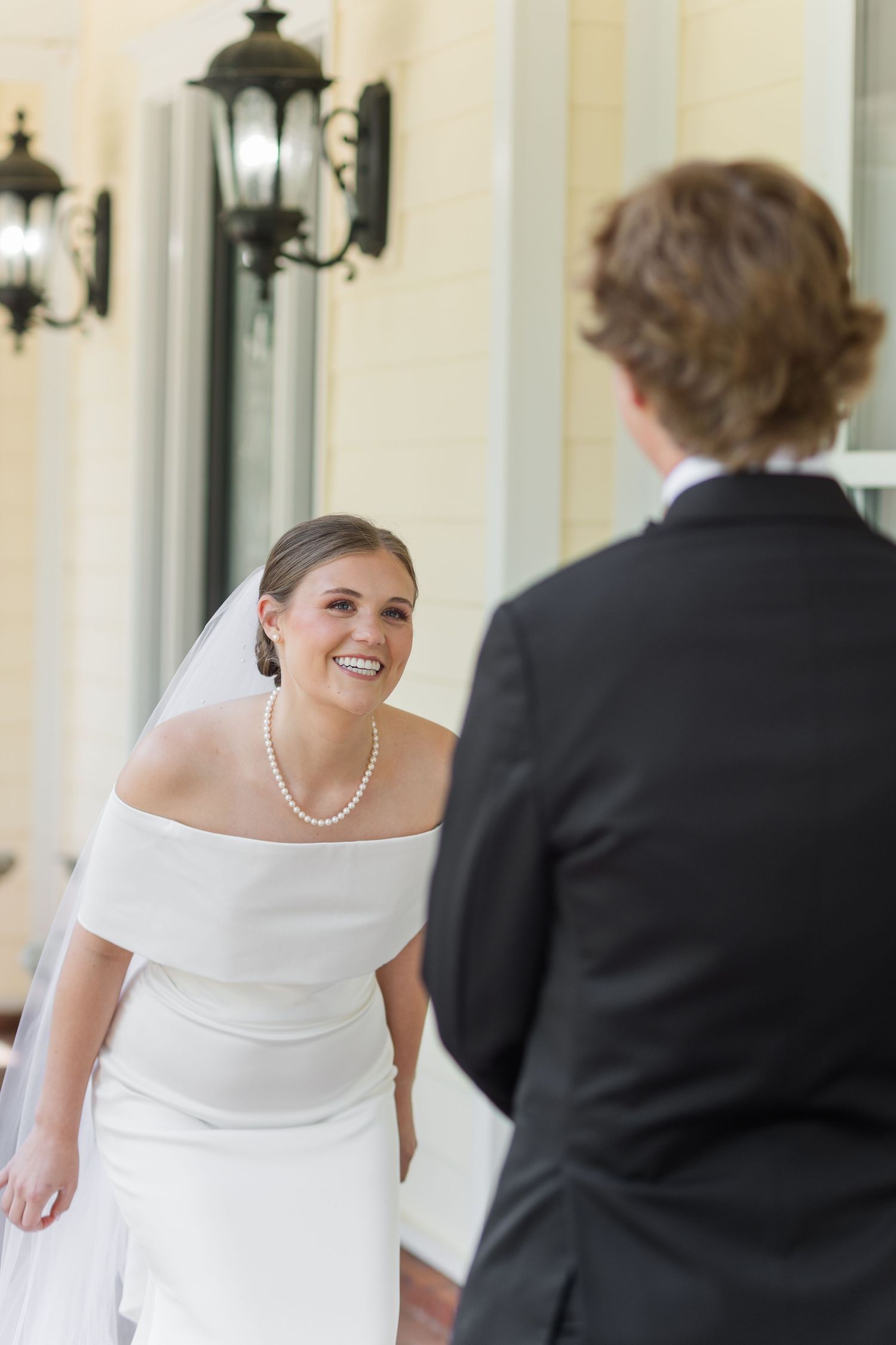 Smiling bride in off-shoulder white gown and pearl necklace shares a first look moment with groom in black suit.