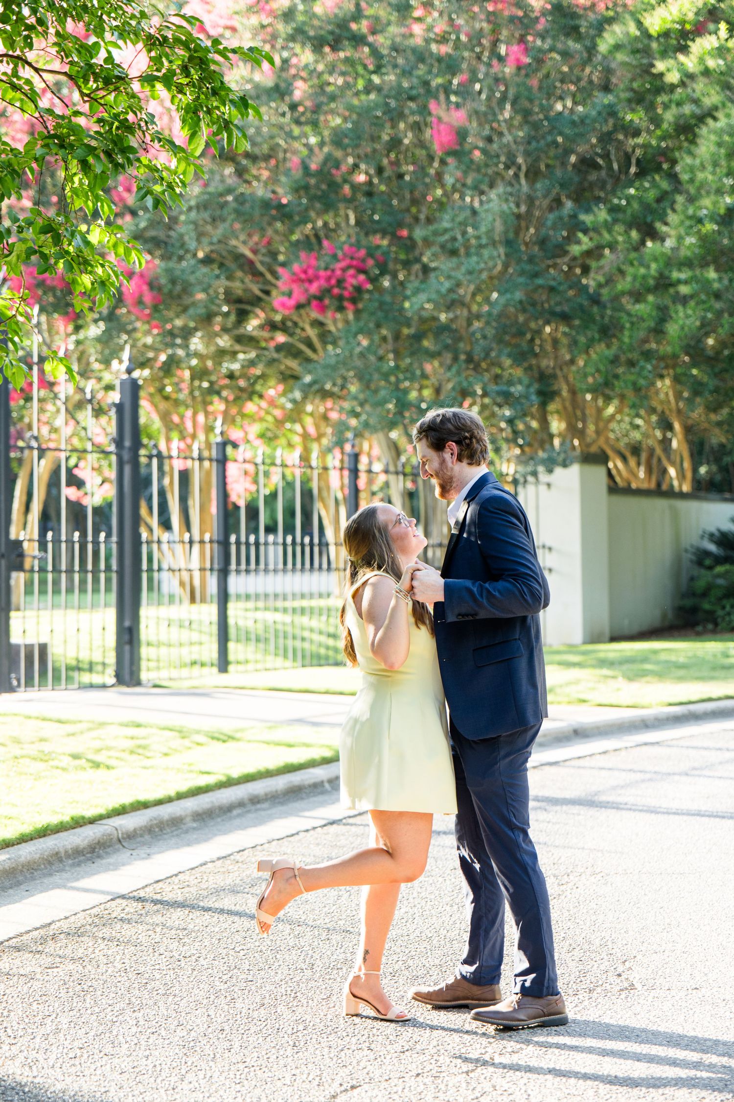 Couple dancing romantically on a sunny street, woman in yellow dress lifting her leg, man in navy suit, pink blooming trees behind.