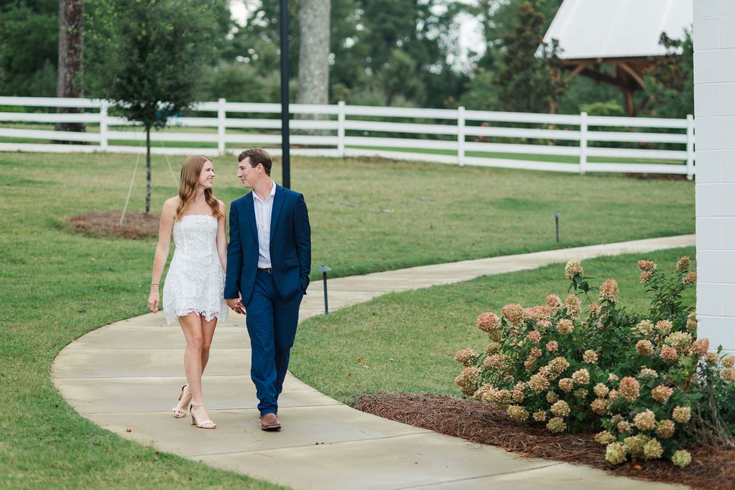 Couple walking hand-in-hand on a curved pathway surrounded by lush greenery and a white fence.