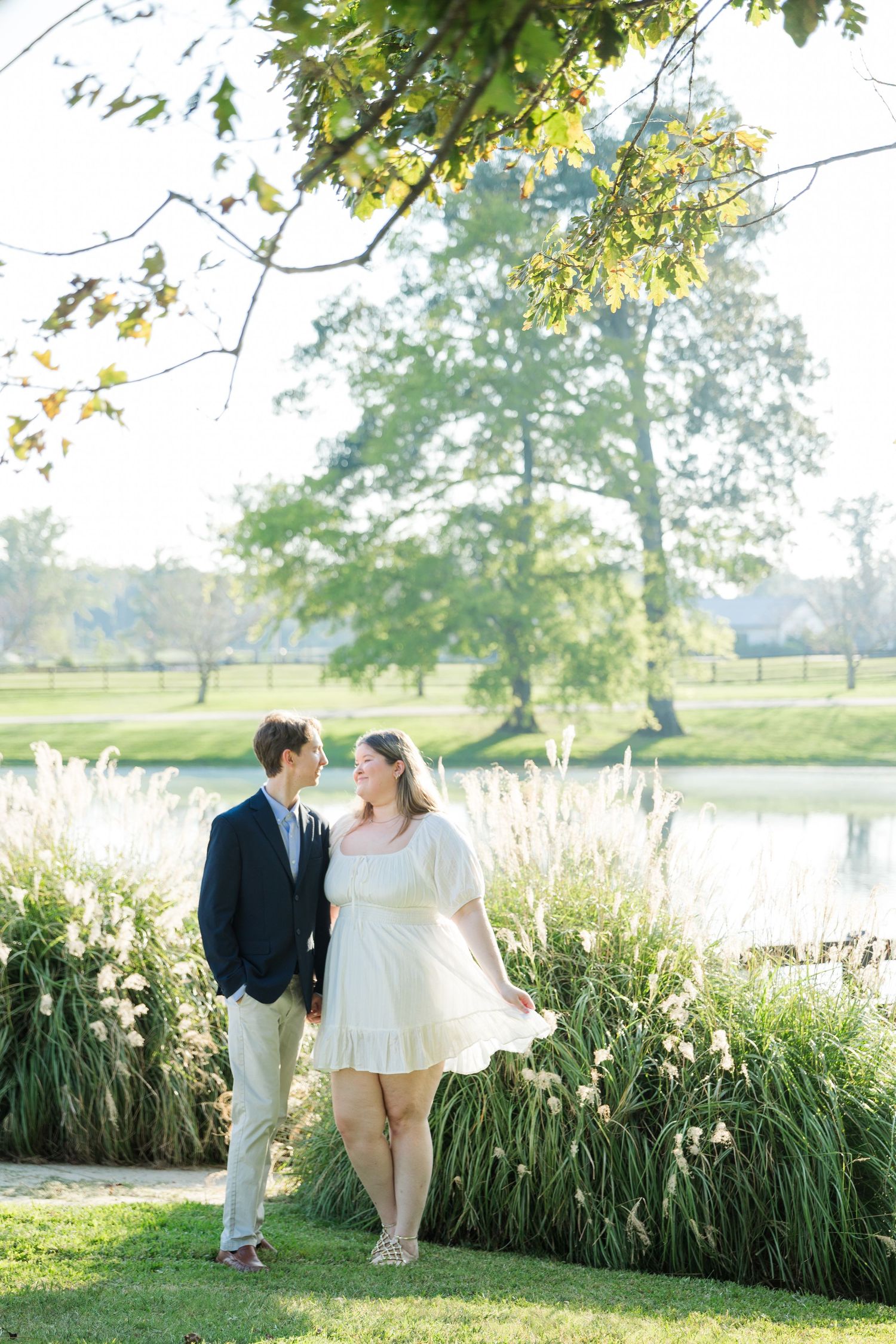 Couple holding hands near ornamental grasses by a pond, man in navy blazer, woman in white dress, sunny outdoor setting.