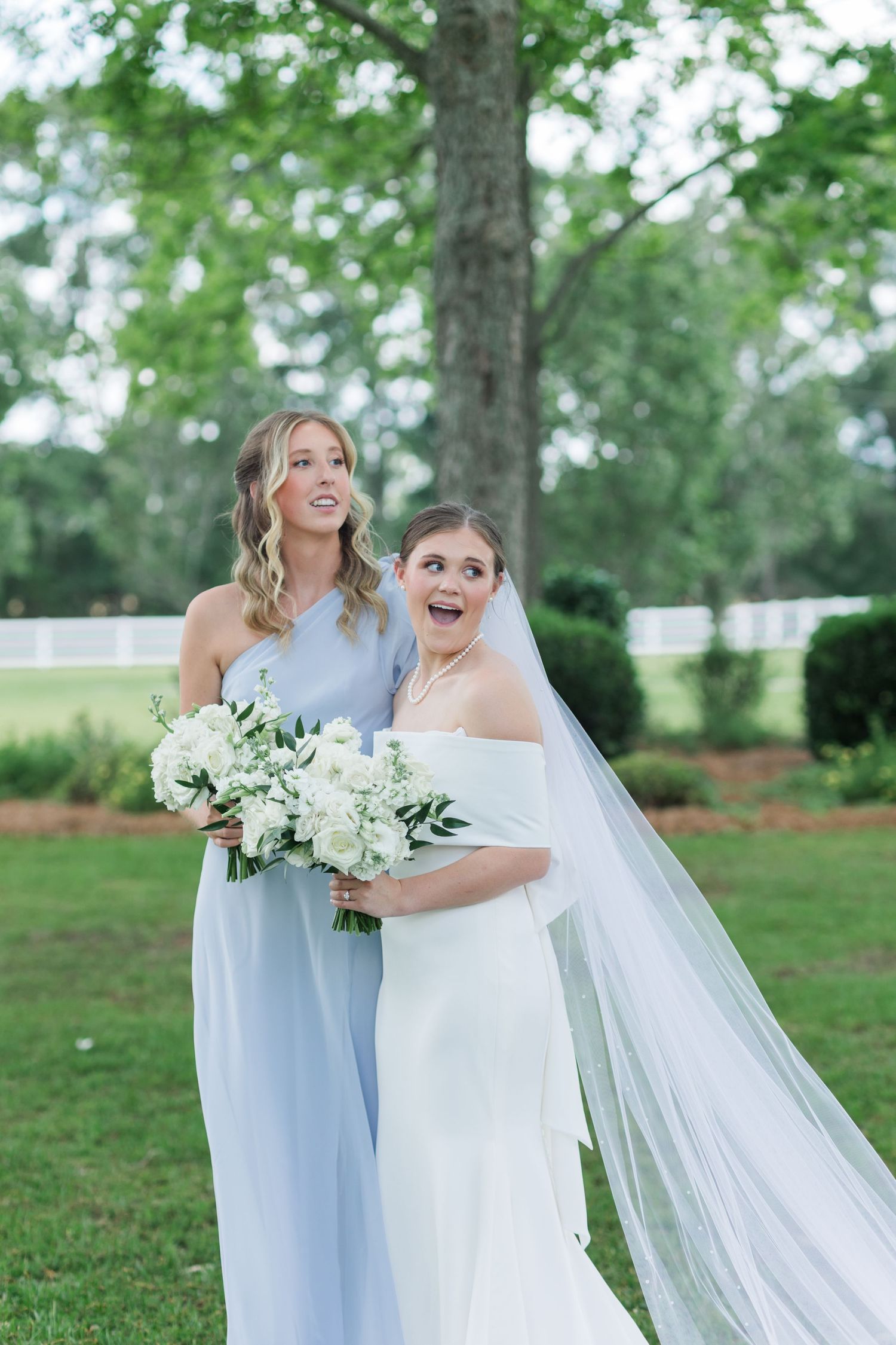 Bride and bridesmaid laugh joyfully outdoors, bride wearing white gown with long veil holding white floral bouquet.