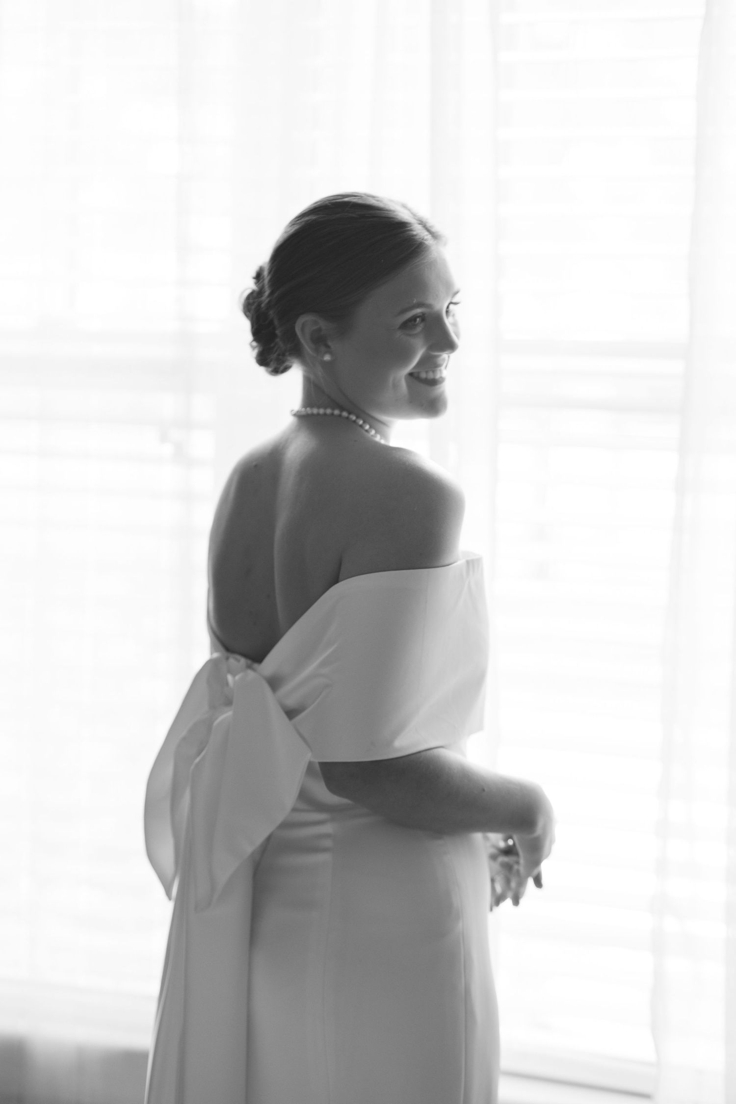 Black and white photo of a smiling bride in an off-shoulder gown with a bow, wearing pearl jewelry, standing by a bright window.