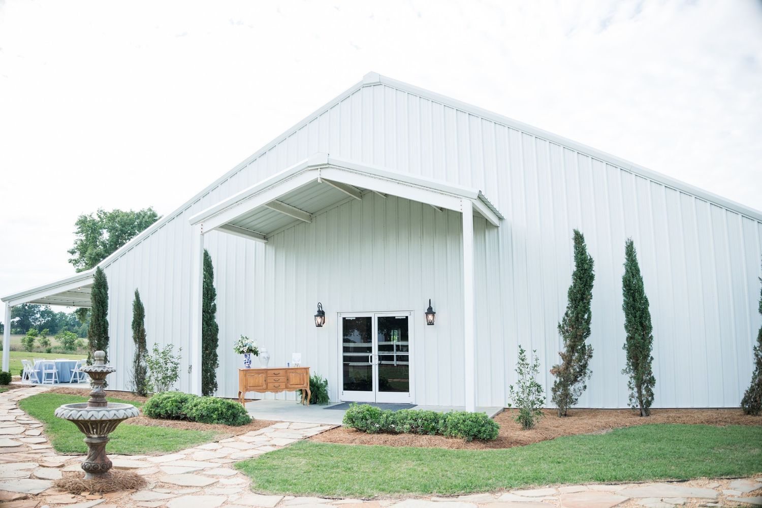 White modern barn wedding venue exterior with cypress trees and stone pathway.