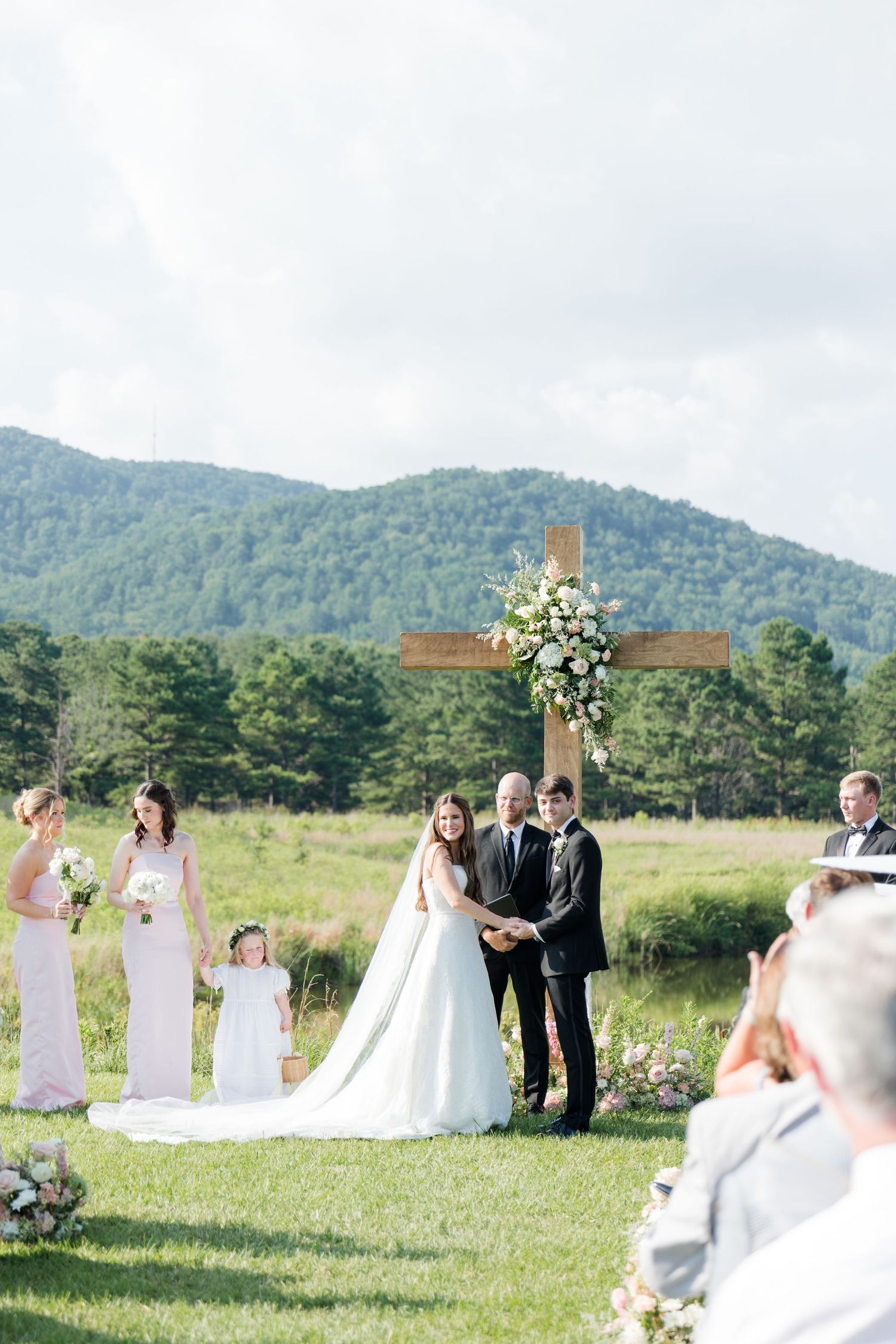 Outdoor wedding ceremony with couple at wooden cross altar against scenic mountain backdrop.