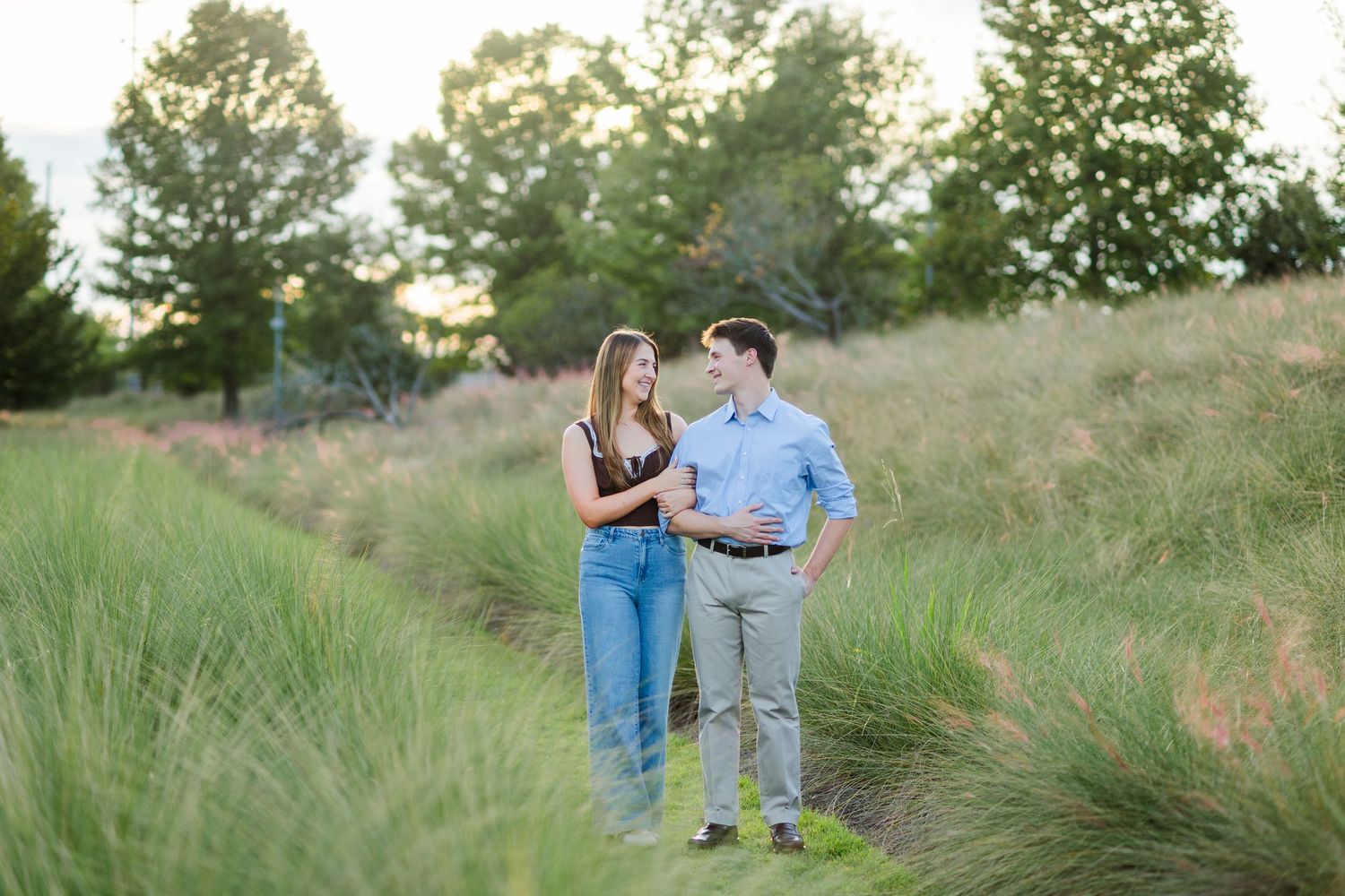 A couple walks hand in hand through a scenic green meadow path at golden hour.