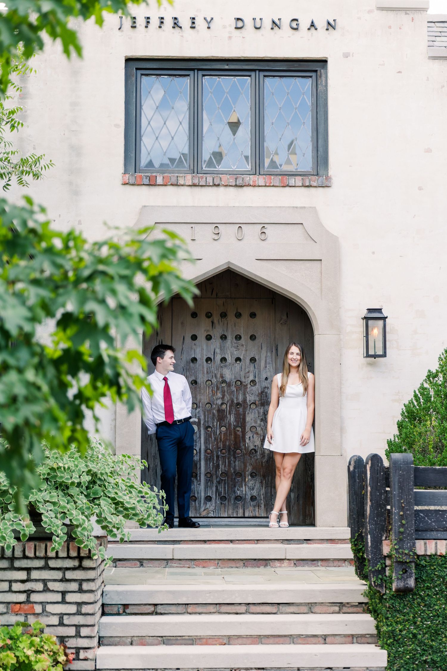 Couple standing in arched entrance of historic Jeffrey Dungan 1906 building surrounded by green foliage.