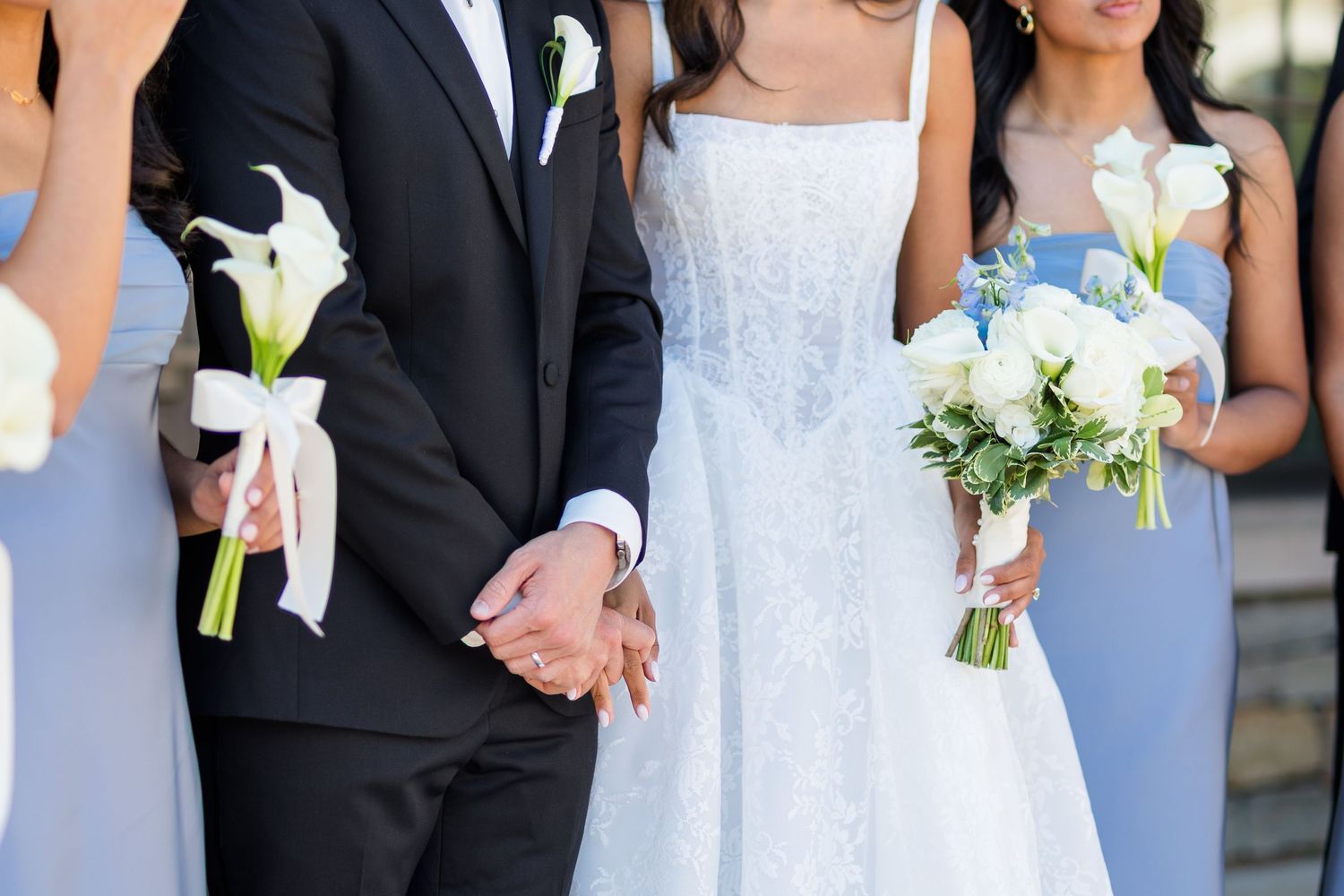 Bride and groom holding hands at wedding ceremony with bridesmaids in blue dresses holding white calla lily bouquets.