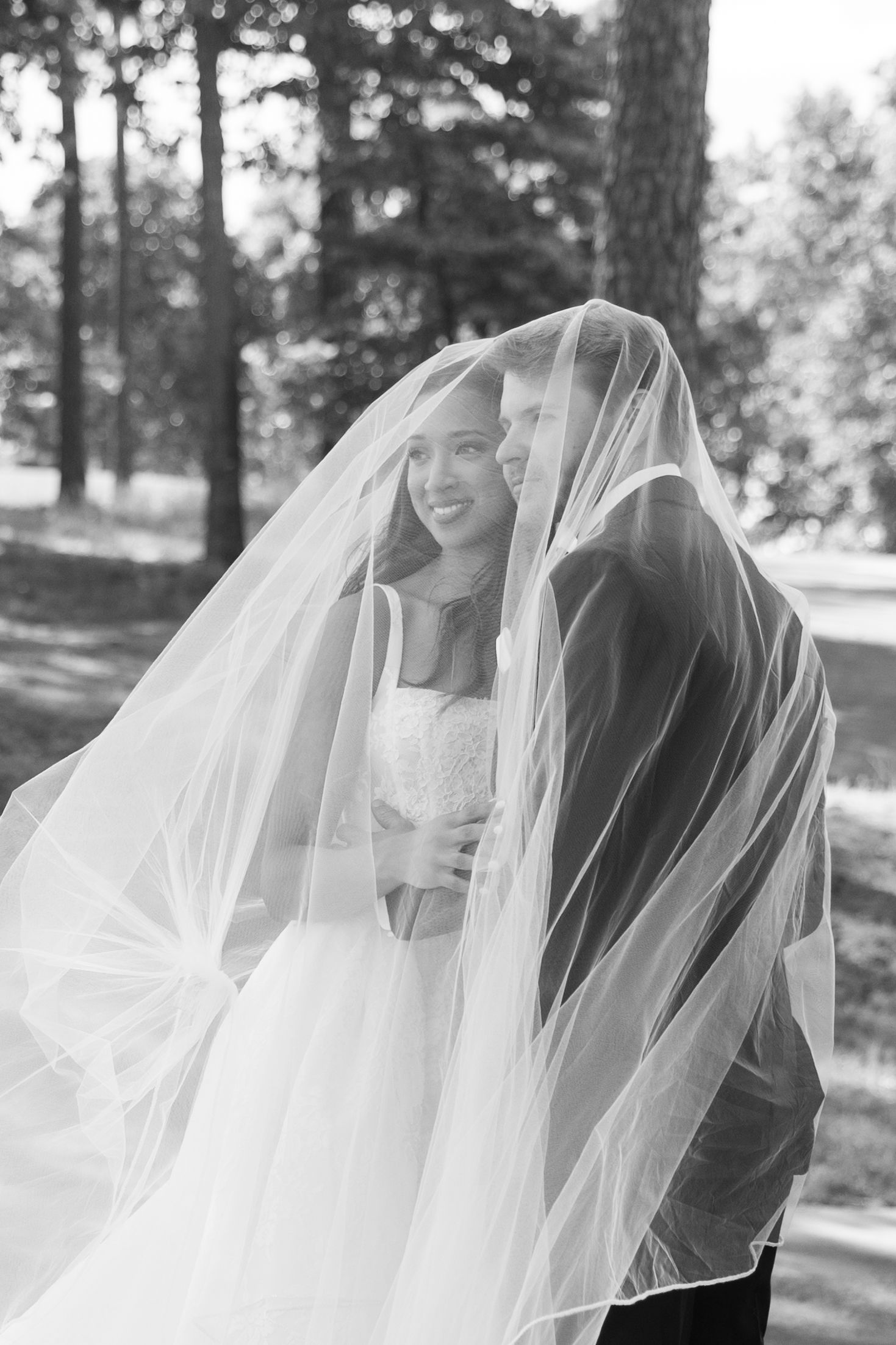 Black and white photo of bride and groom sharing a romantic moment under a flowing wedding veil outdoors.