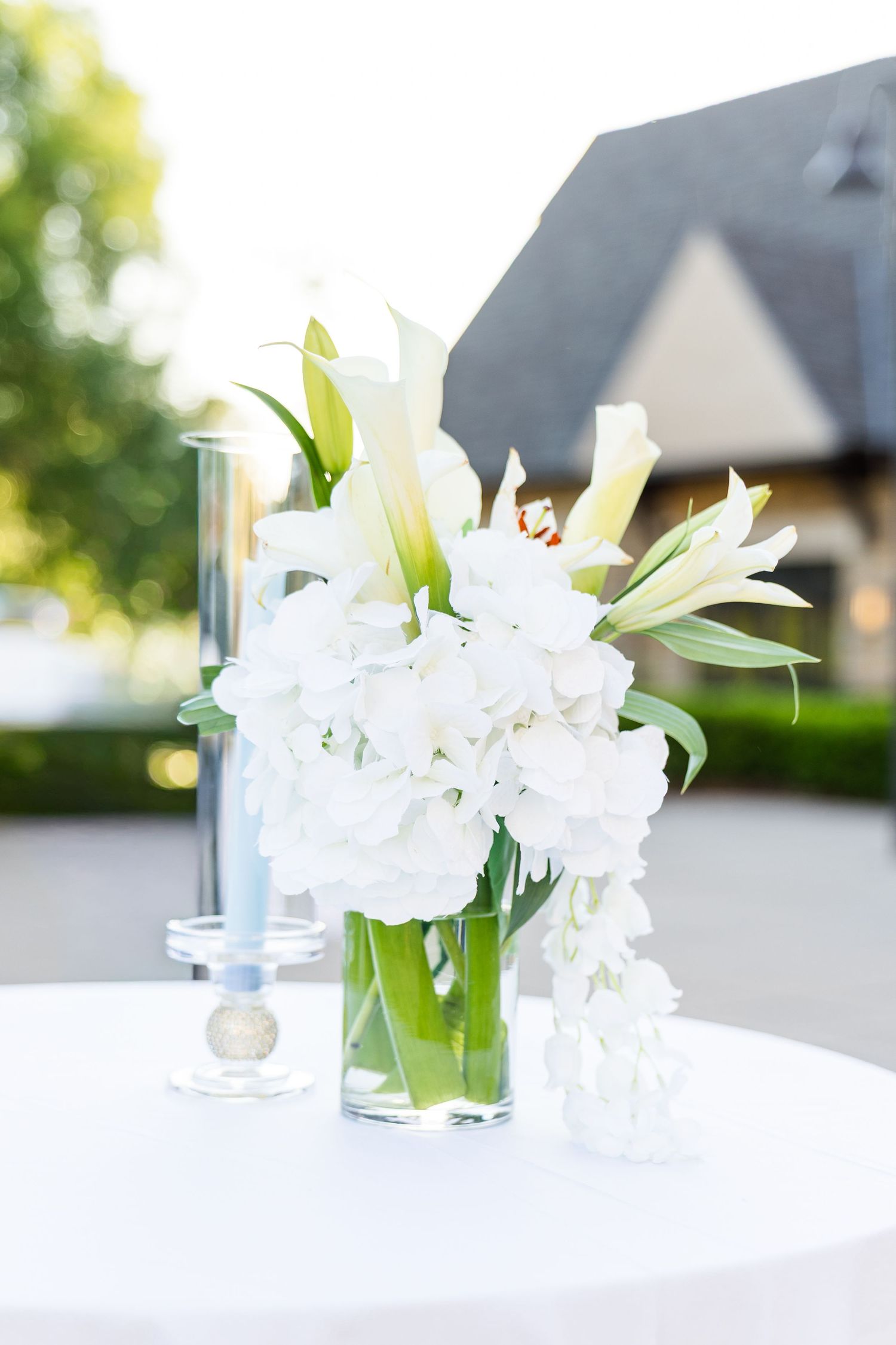 Elegant white floral centerpiece with lilies and hydrangeas in a glass vase on a white tablecloth at an outdoor event.