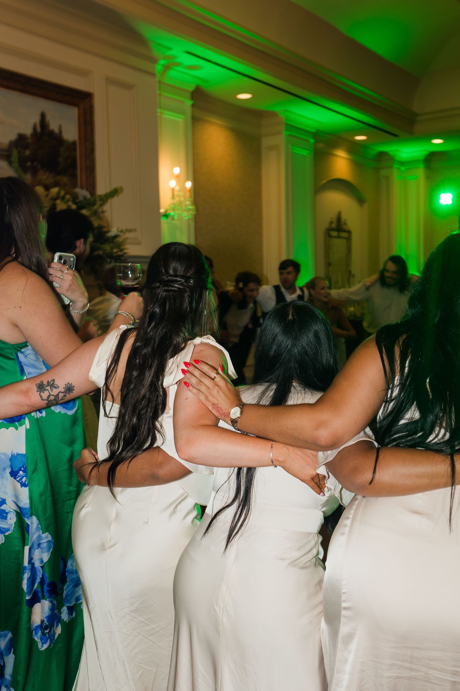 Bridesmaids in white dresses dancing together under green lights at lively wedding reception party.