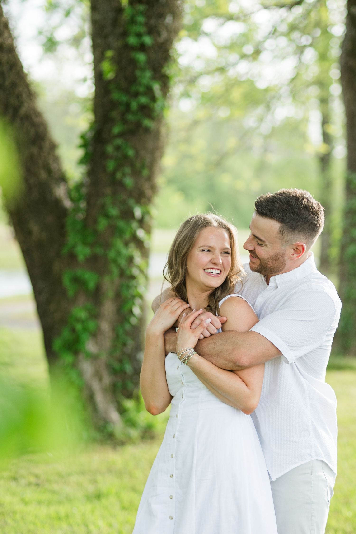 Man embracing woman in white dress while smiling in sunlit park with ivy-covered trees behind them.