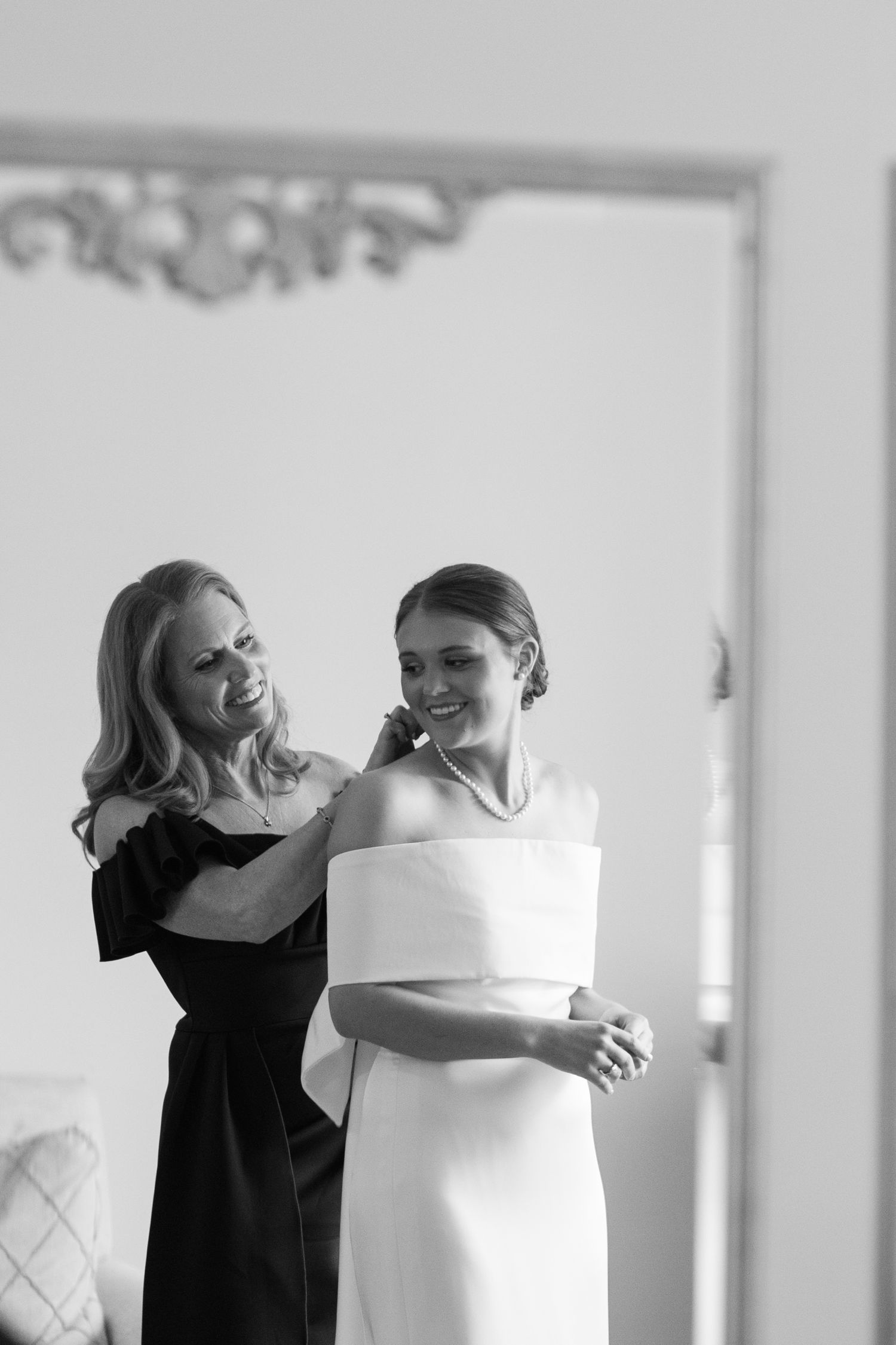 Black and white photo of a bride in white gown smiling as a woman in black dress fastens her necklace.