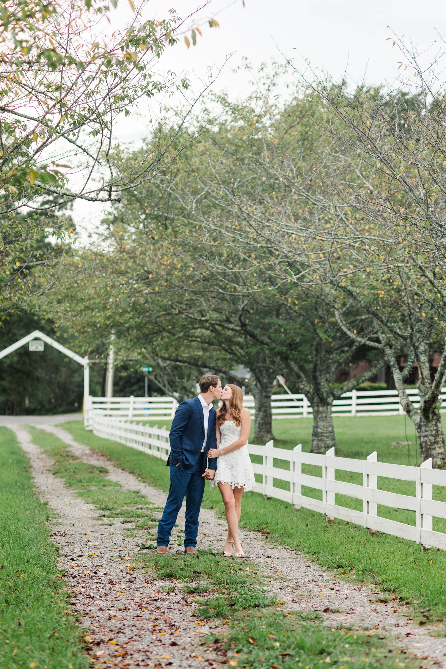 Couple sharing a kiss on a tree-lined gravel path beside a white fence at a rustic farm venue.