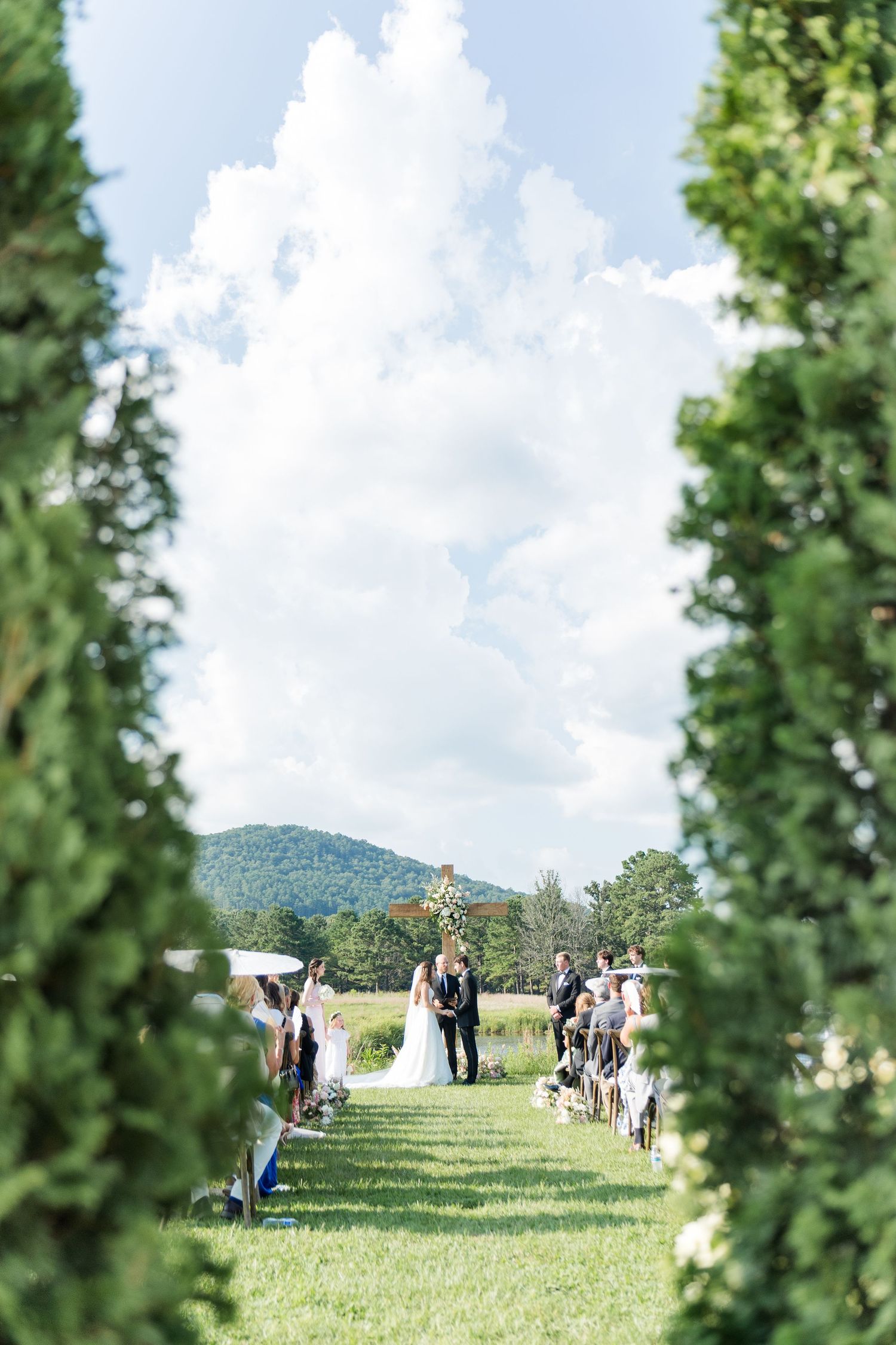 Outdoor wedding ceremony on green lawn framed by tall trees with mountain backdrop and dramatic cloudy sky.