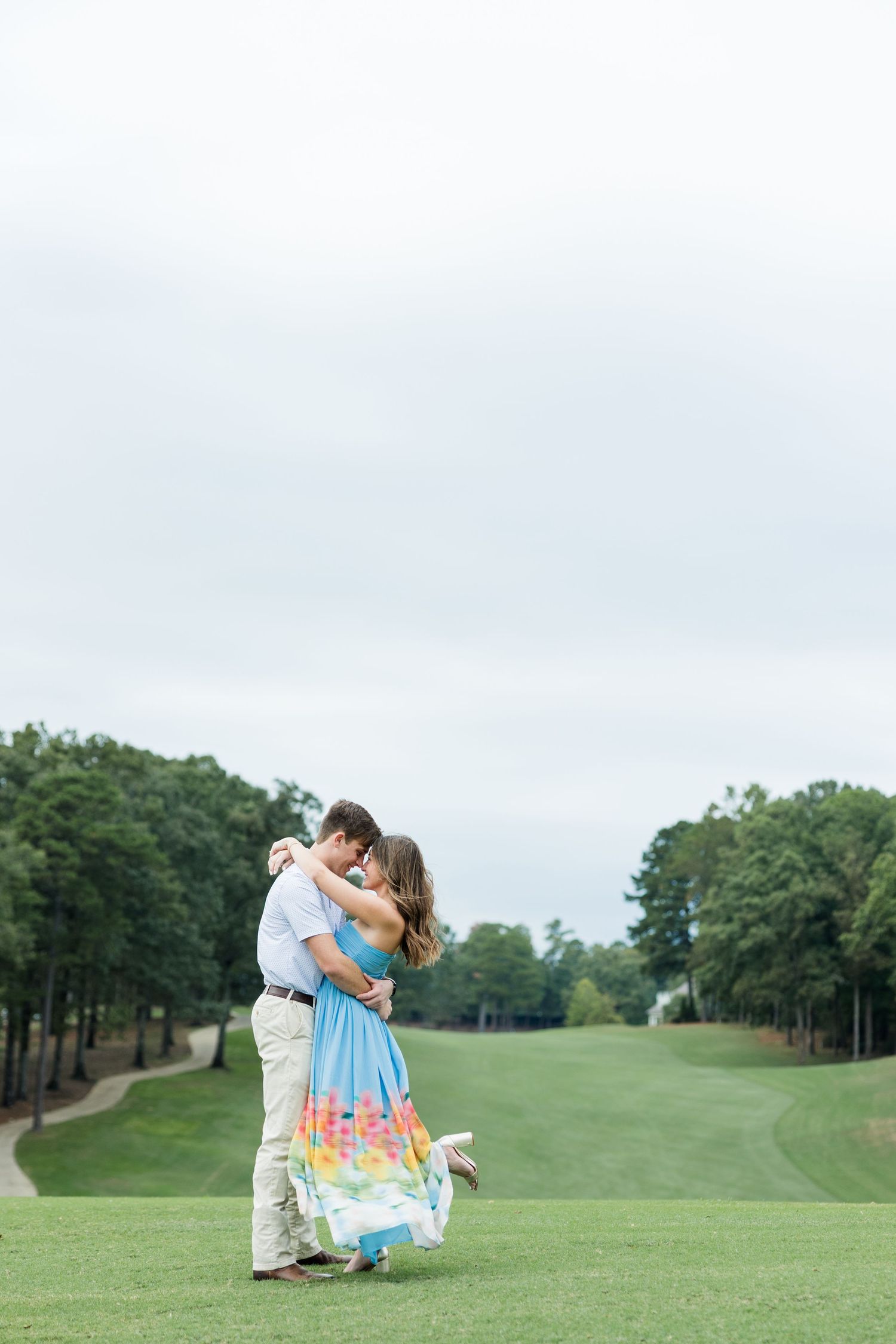 Couple embracing on a golf course fairway as he lifts her in a colorful floral dress, surrounded by trees.