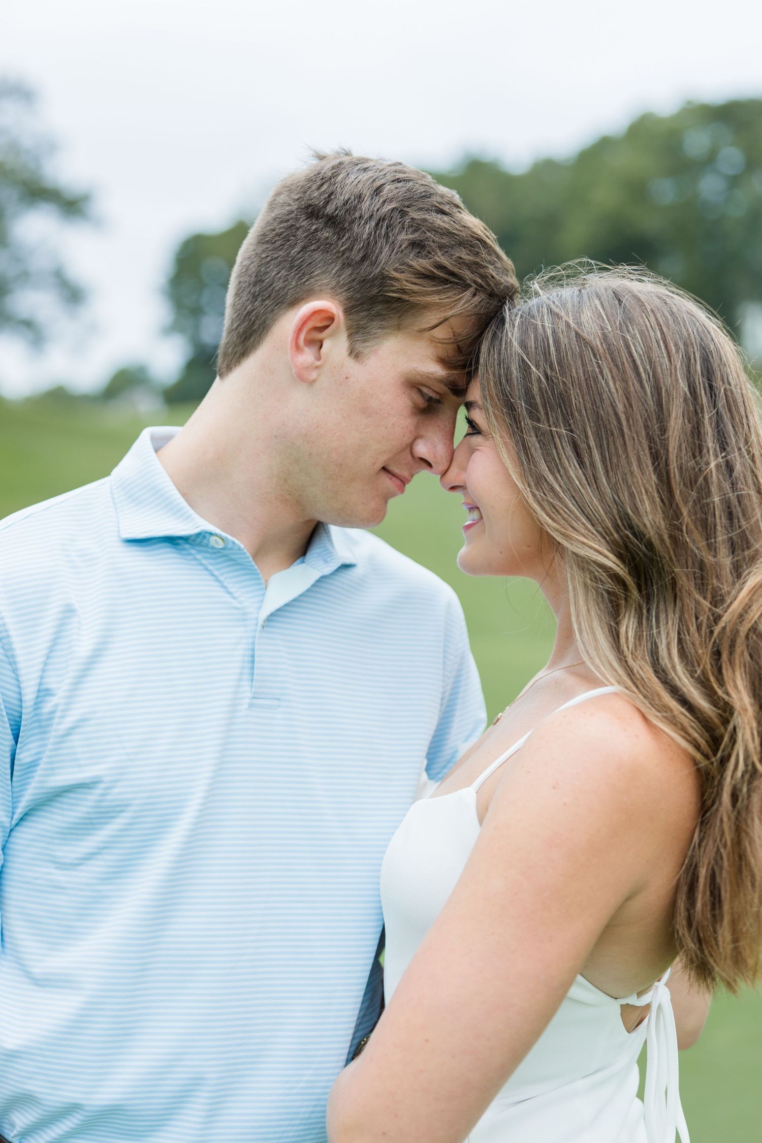 Couple touching forereads with eyes closed and smiling softly on a green lawn, she in a white dress and he in a light blue polo shirt.