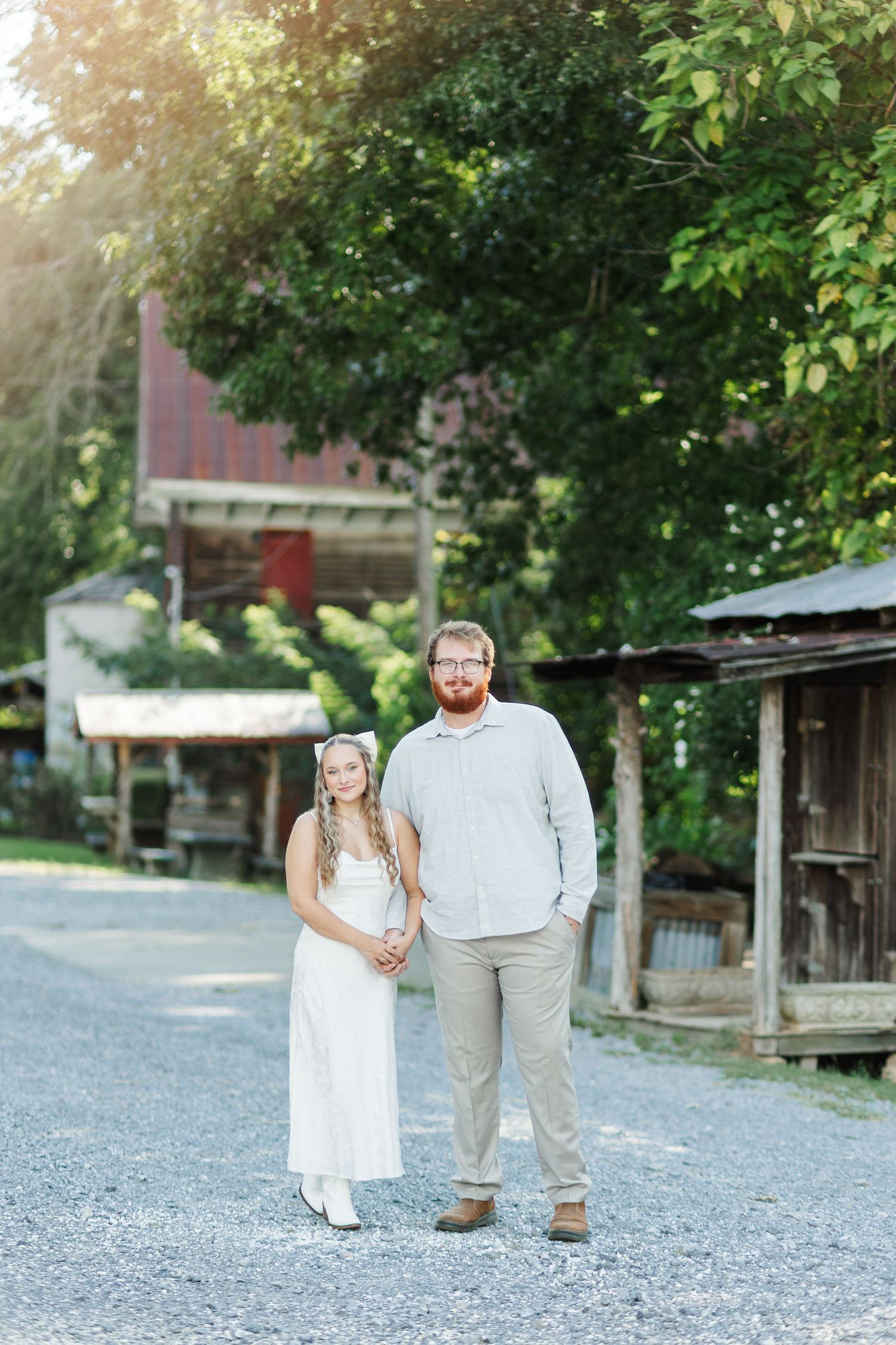 Bride and groom stand together on gravel path surrounded by trees near rustic buildings.