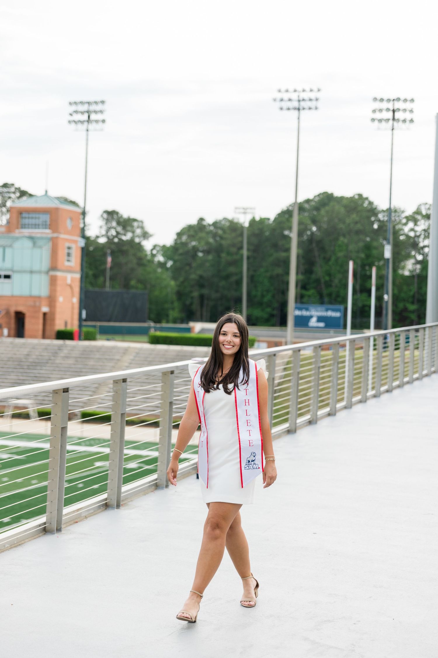 Young woman in white dress and graduation stole walks along stadium walkway during outdoor graduation photo session.