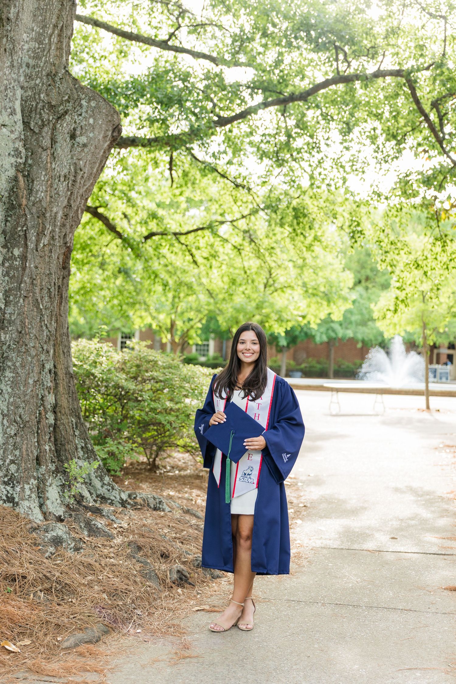 Young woman in navy blue graduation gown smiling outdoors on campus path surrounded by lush green trees.