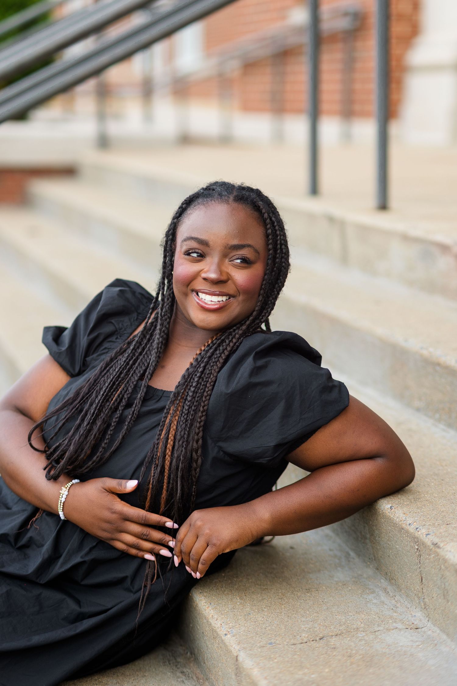 Smiling young Black woman with long braids wearing a black dress, sitting on outdoor steps.