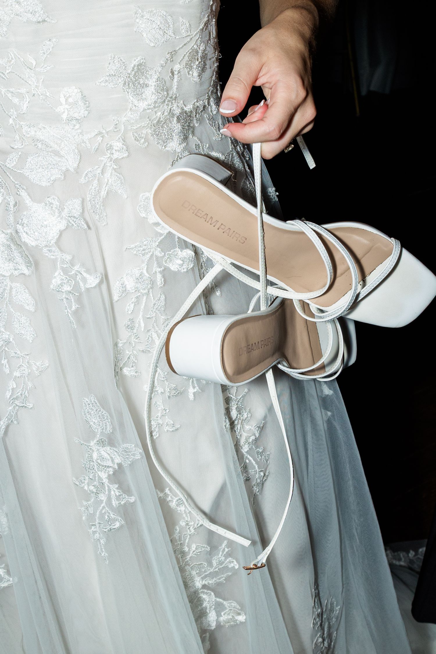 Bride holding white strappy heeled sandals against her lace embroidered wedding gown.
