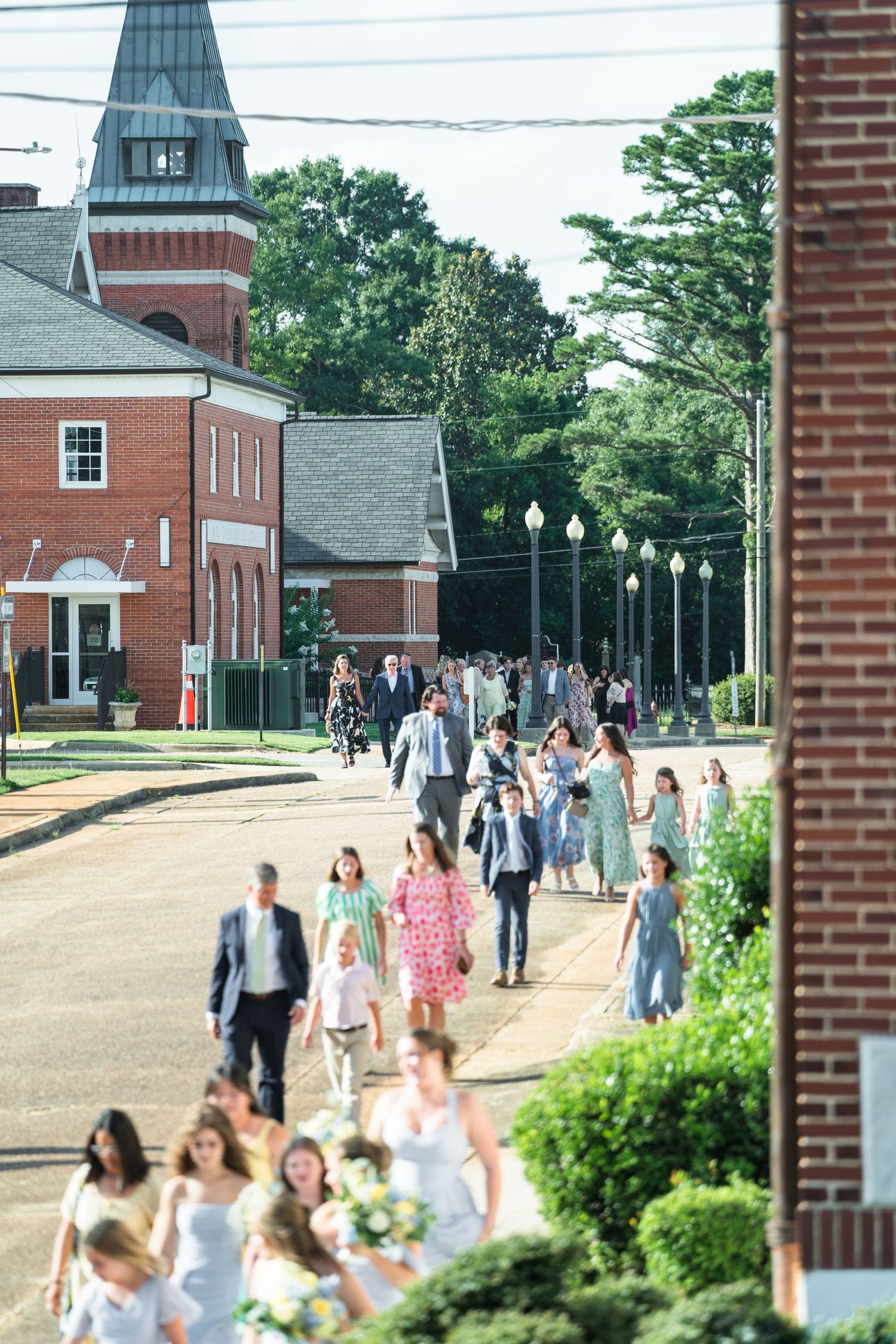 Wedding guests and bridal party walk down sunny small-town street after outdoor church ceremony in summer.