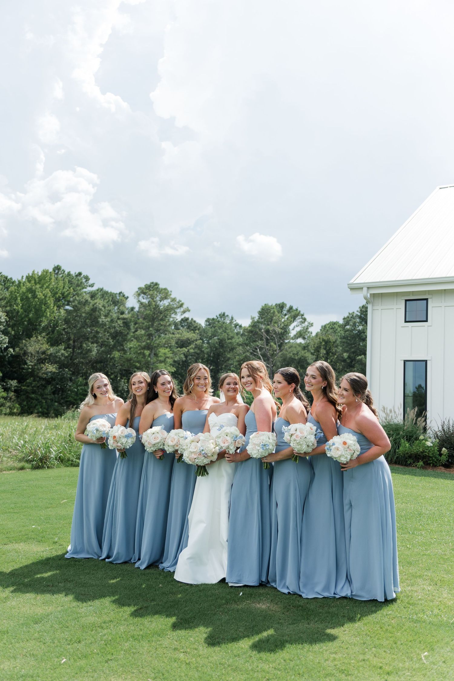 Bride in white gown with bridesmaids in dusty blue dresses holding white floral bouquets on green lawn.