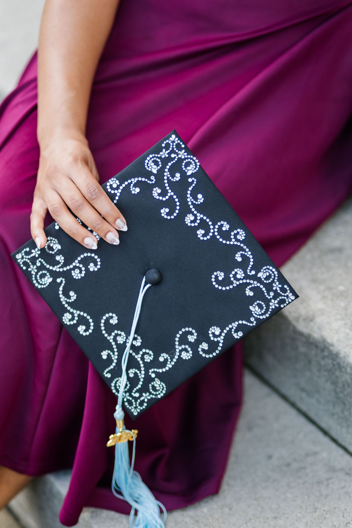 Close-up of decorated black graduation cap with white swirl designs and blue tassel on burgundy dress.