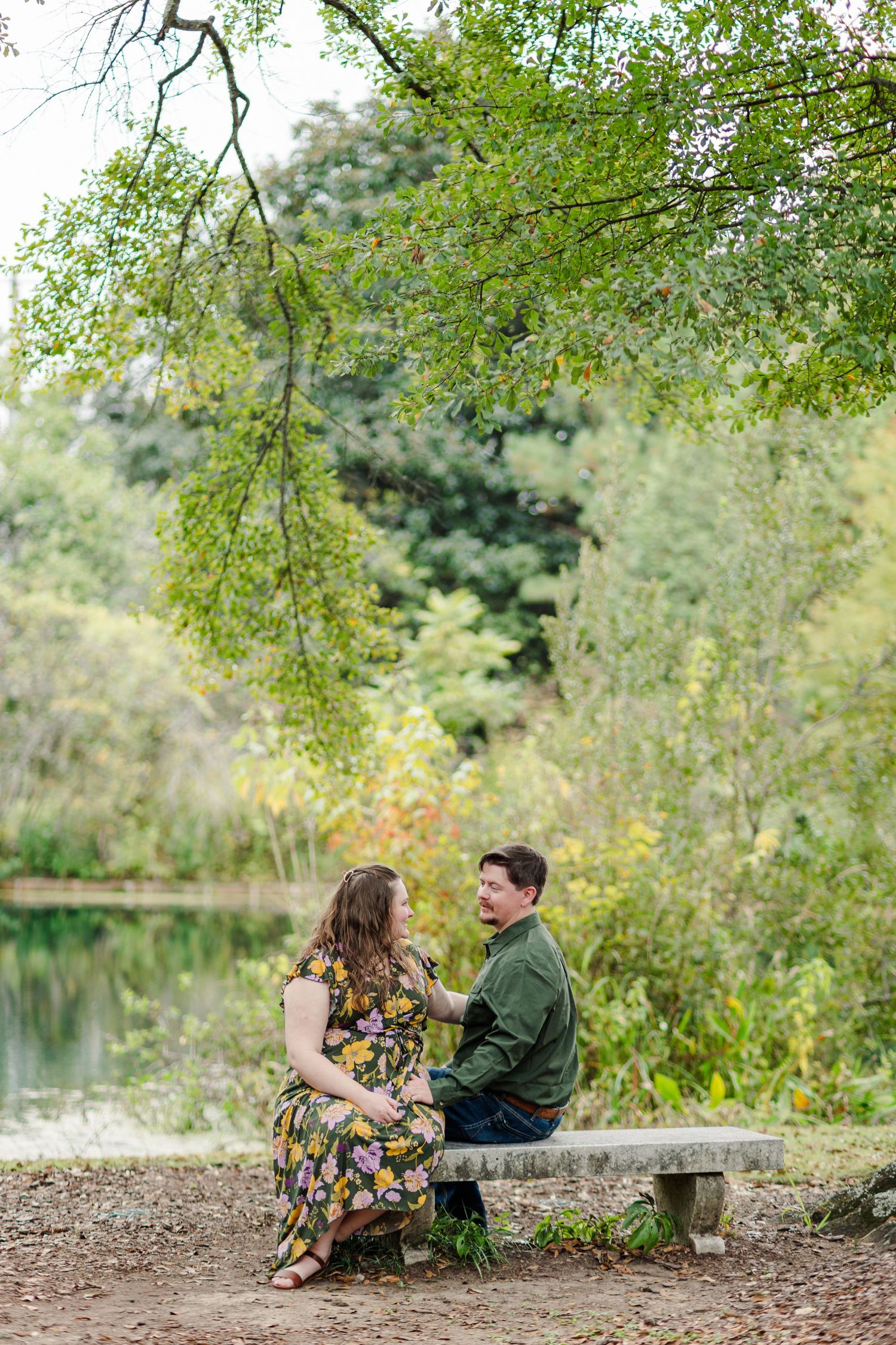 Couple sitting on a stone bench by a pond, surrounded by lush green trees, sharing a joyful moment outdoors.