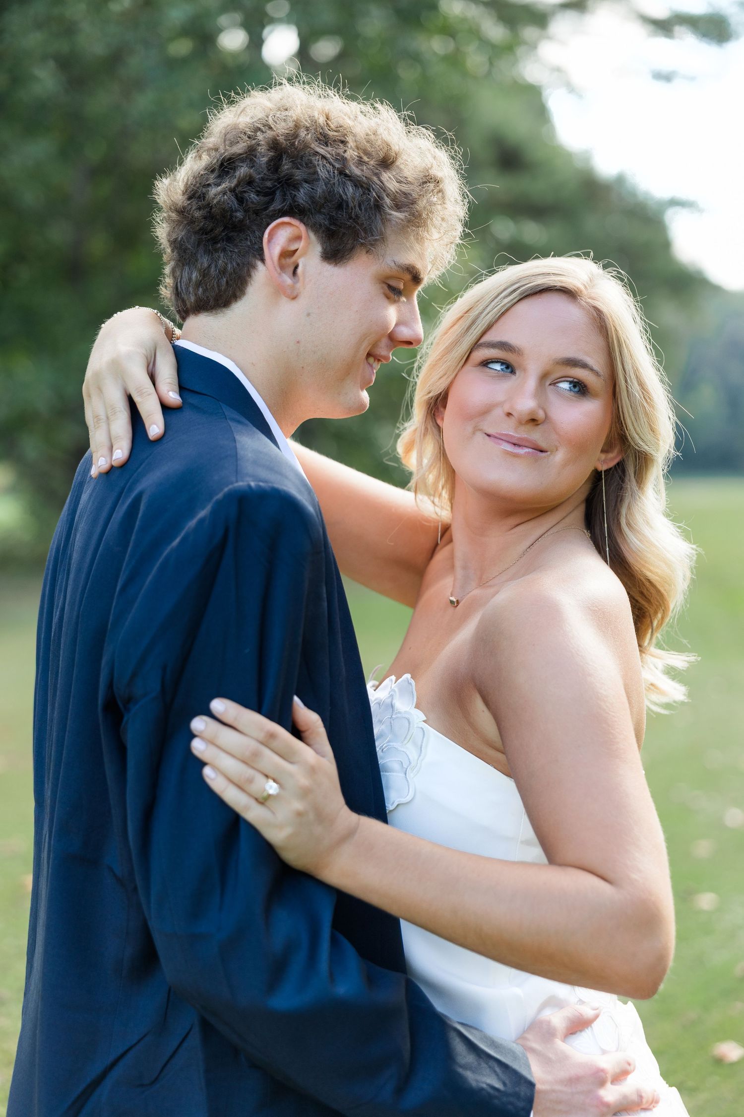 A bride and groom embrace outdoors, she smiles wearing a white dress and engagement ring, he wears a navy suit.