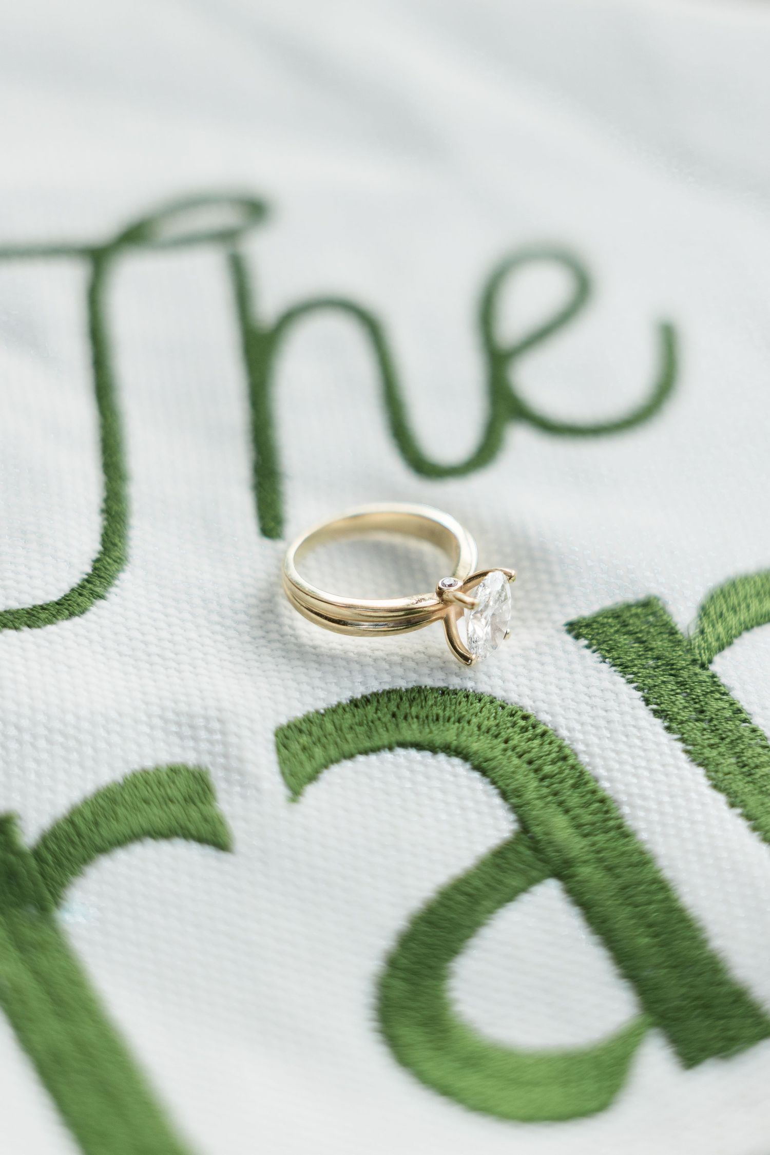 Gold solitaire diamond engagement ring resting on embroidered white fabric with green cursive lettering.