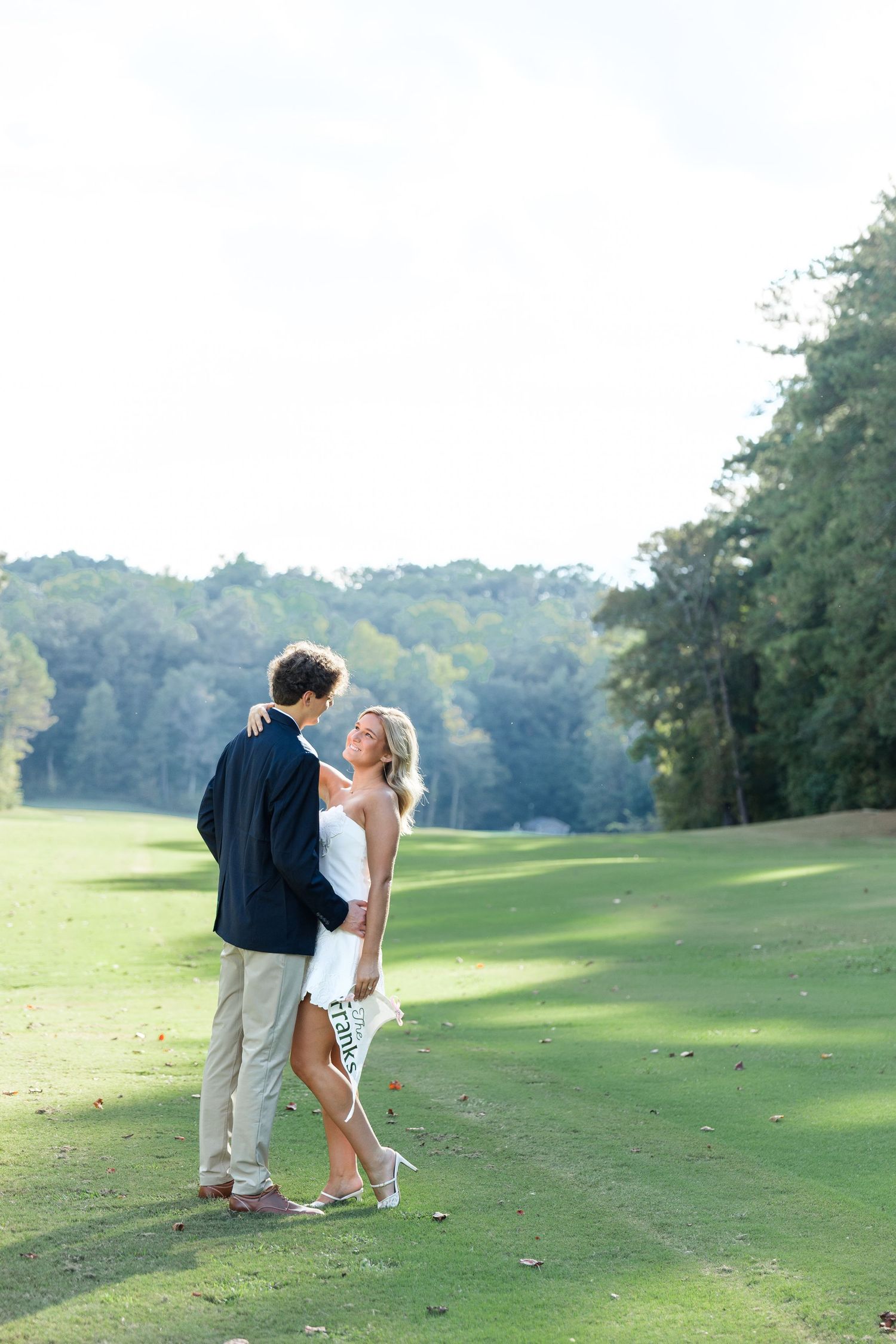 Couple in wedding attire embrace on sunny golf course fairway surrounded by lush green trees.