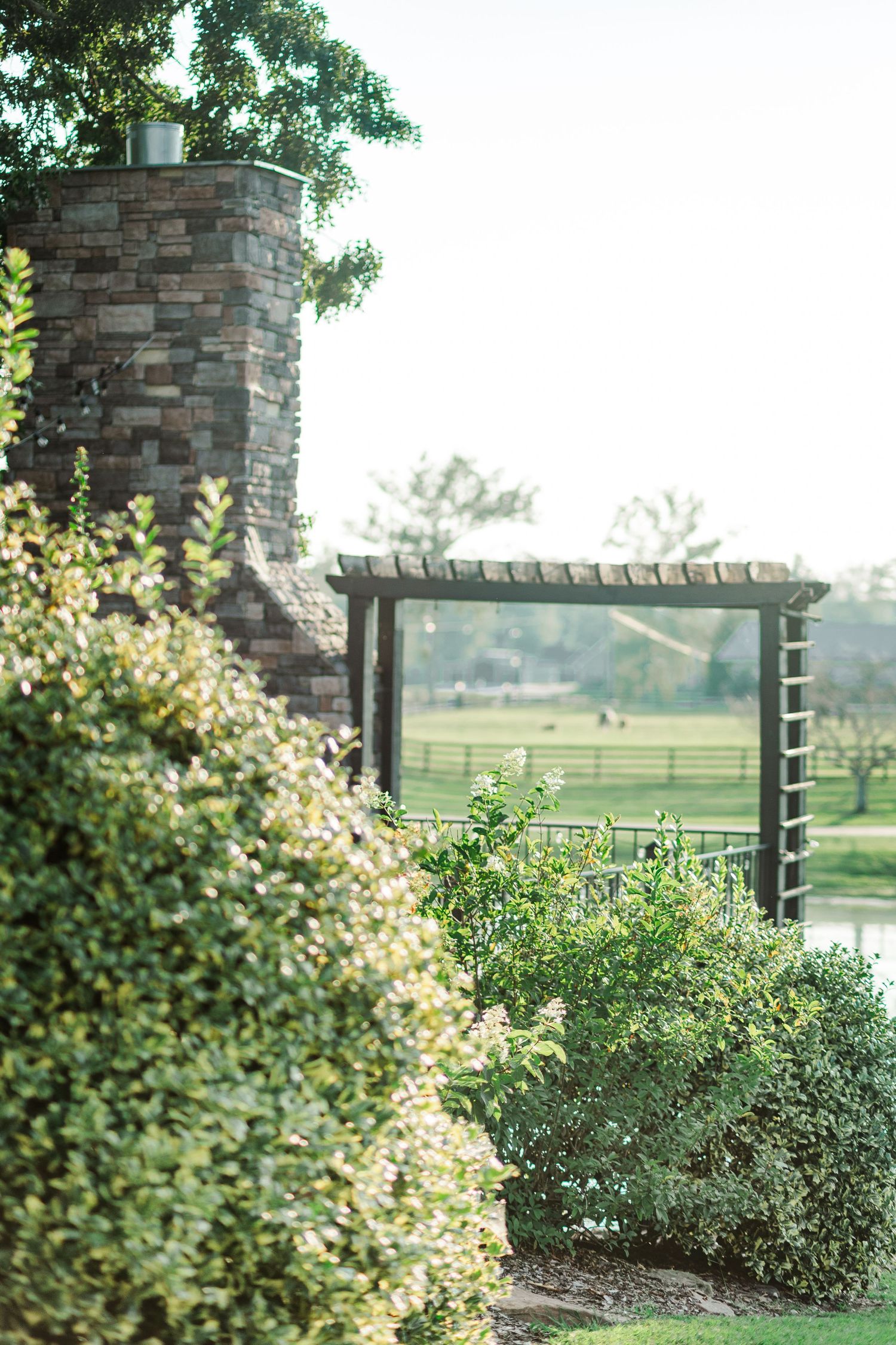 Lush green garden with stone chimney, wooden pergola, and manicured shrubs overlooking a serene open field.