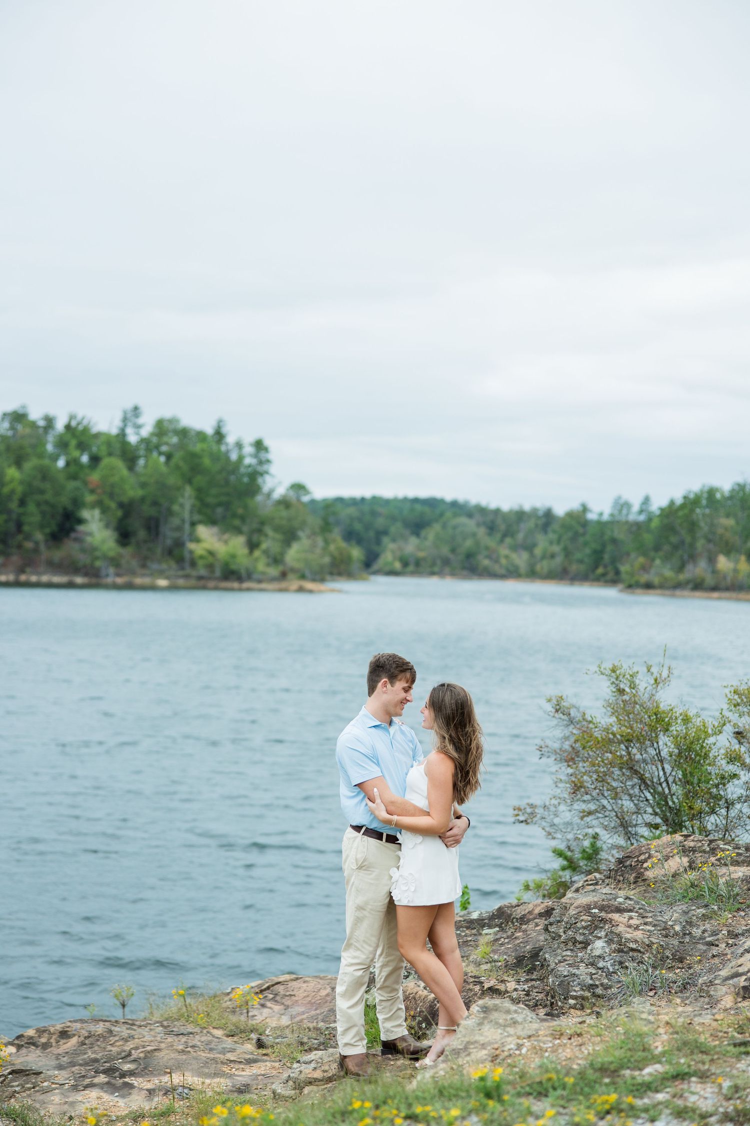 Couple embracing on a rocky shoreline overlooking a calm lake with a tree-lined horizon on a cloudy day.