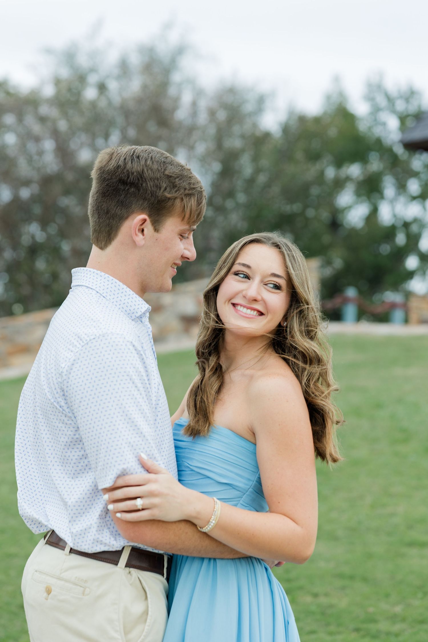 Smiling woman in a blue strapless dress embraces a man in a white shirt and khakis on a green lawn during an engagement photo session.