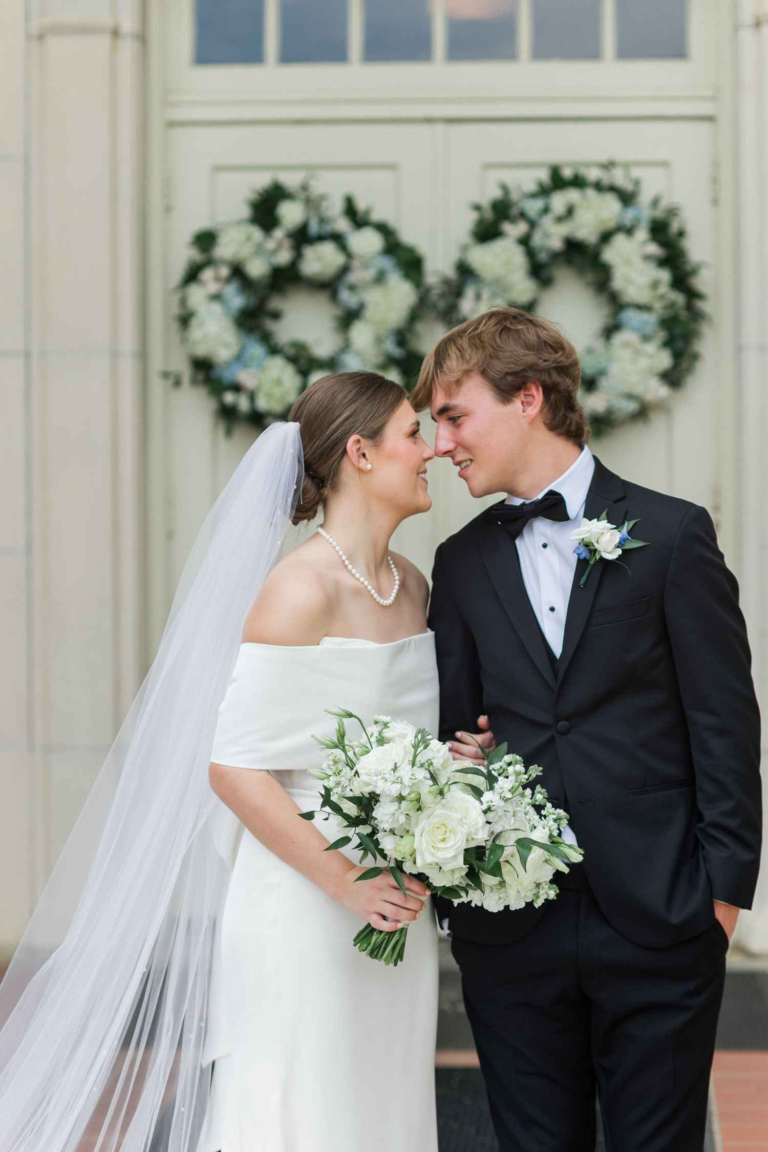 Bride and groom share a romantic moment in front of floral wreaths, she holds a white bouquet in an off-shoulder gown.