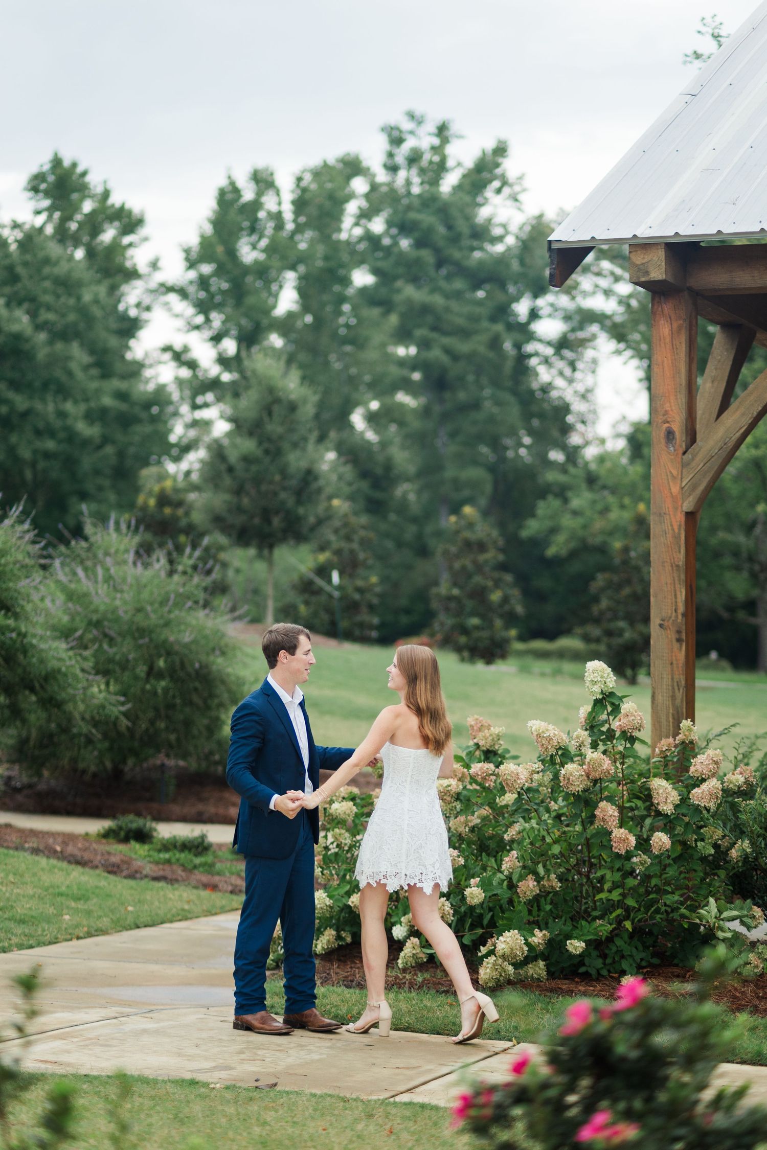 Engaged couple holding hands on garden path near wooden pergola with blooming hydrangea bushes.