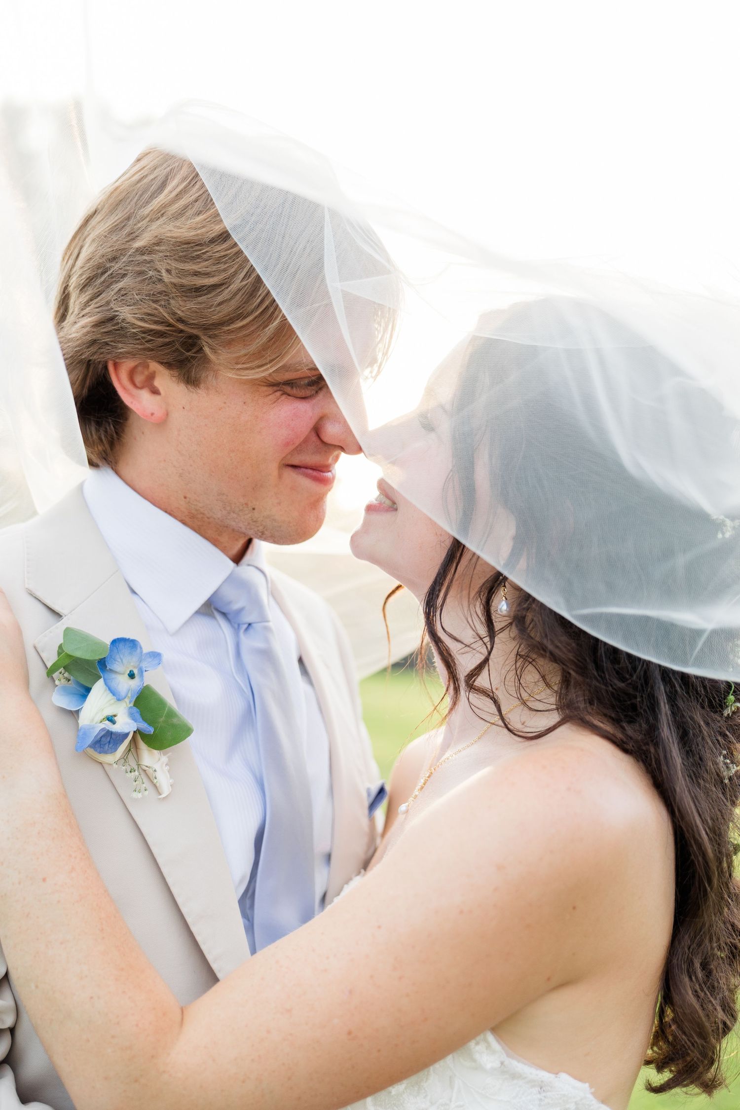 Bride and groom share a romantic moment under a wedding veil, groom wearing a tan suit with blue boutonniere.