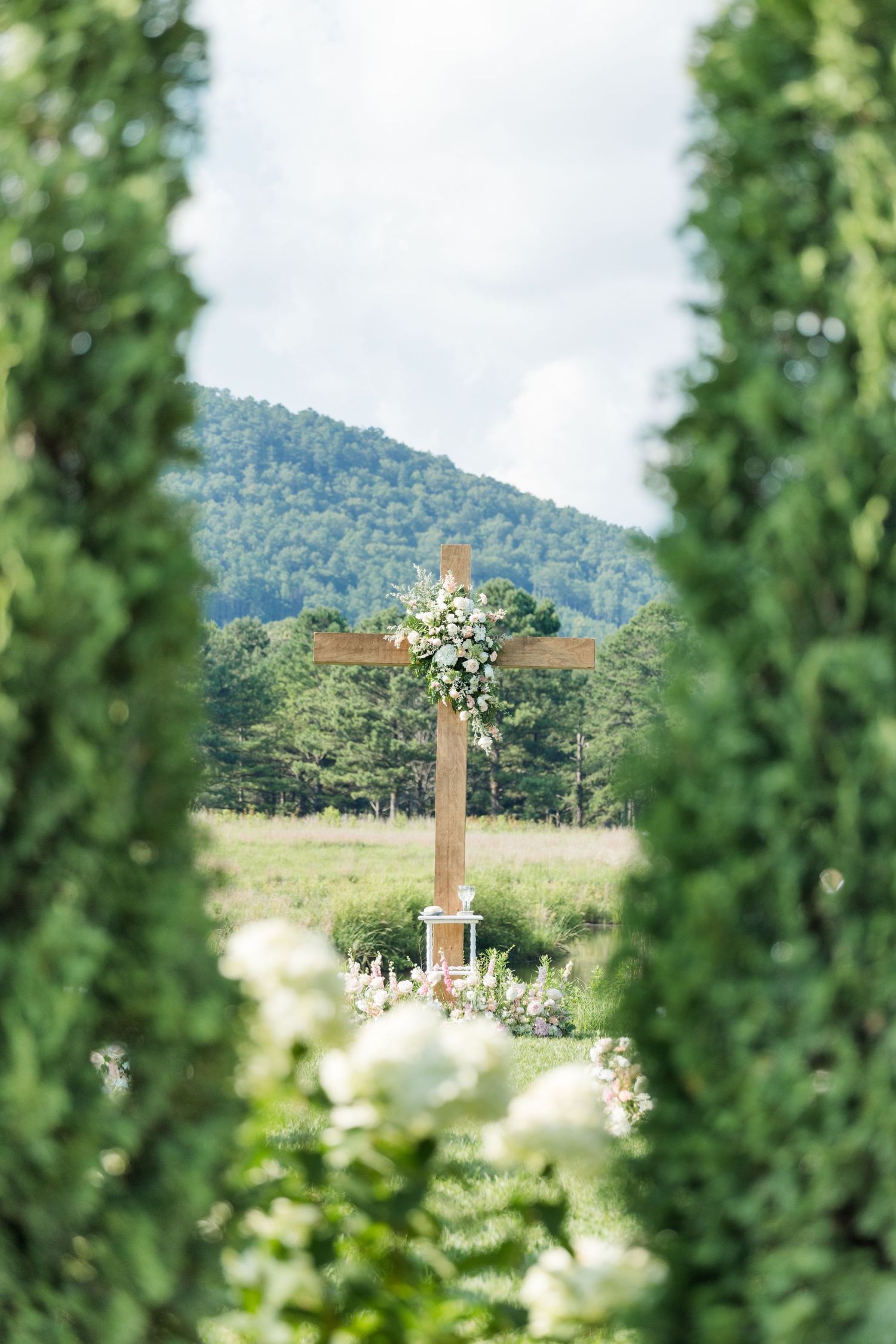 Wooden cross adorned with white flowers framed by tall green cypress trees with mountain backdrop.