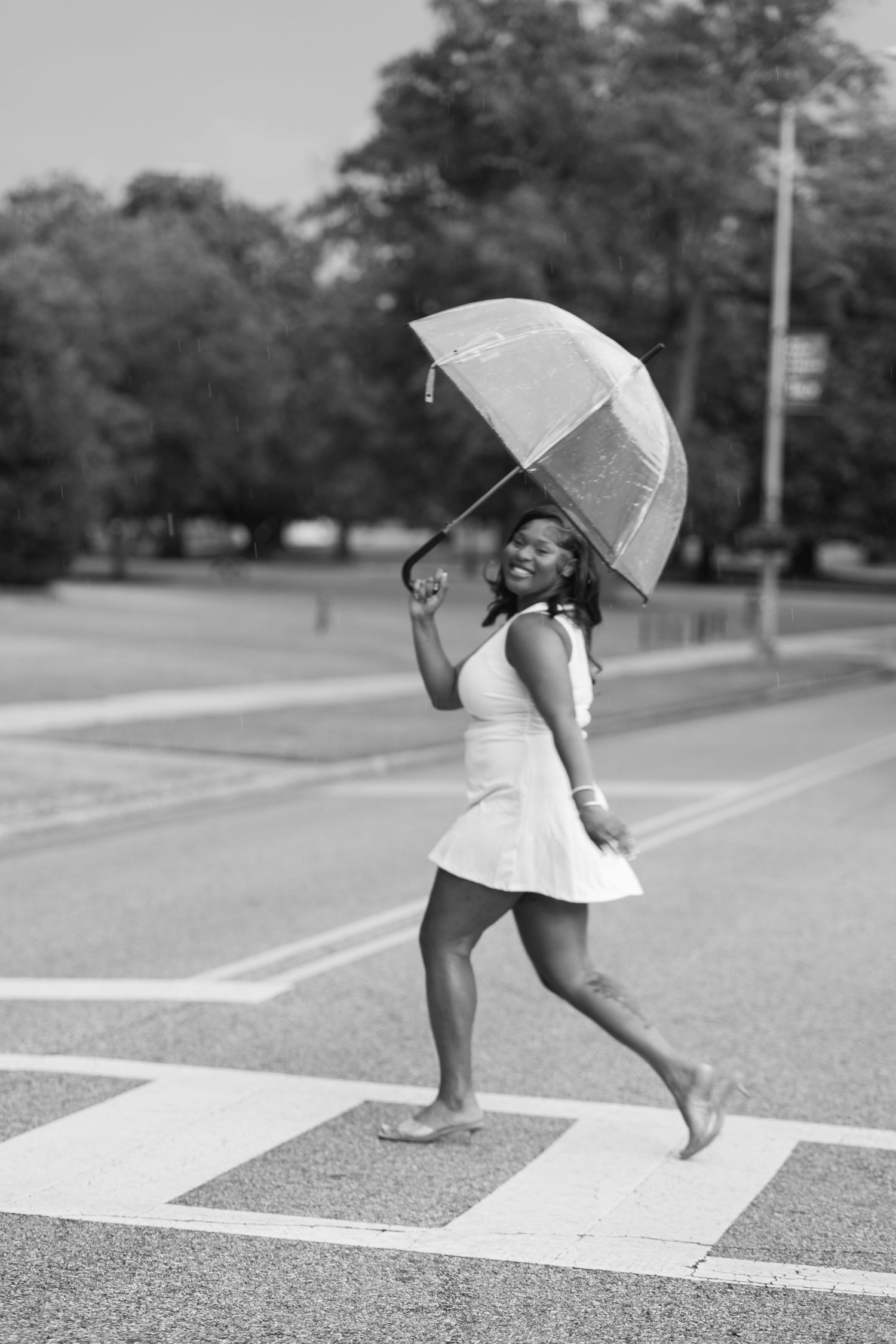 Black and white photo of a smiling woman in a dress holding an umbrella, skipping barefoot across a crosswalk.