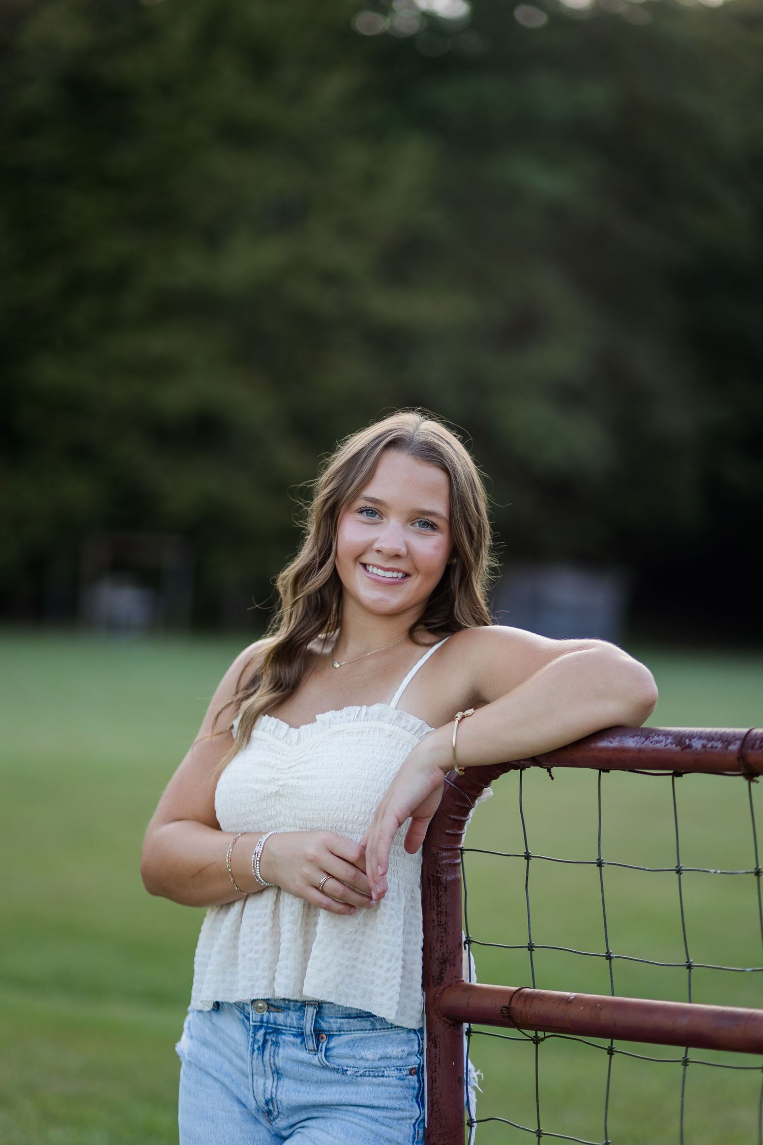 Young woman in white crop top and jeans smiling while leaning on a rustic red gate outdoors.