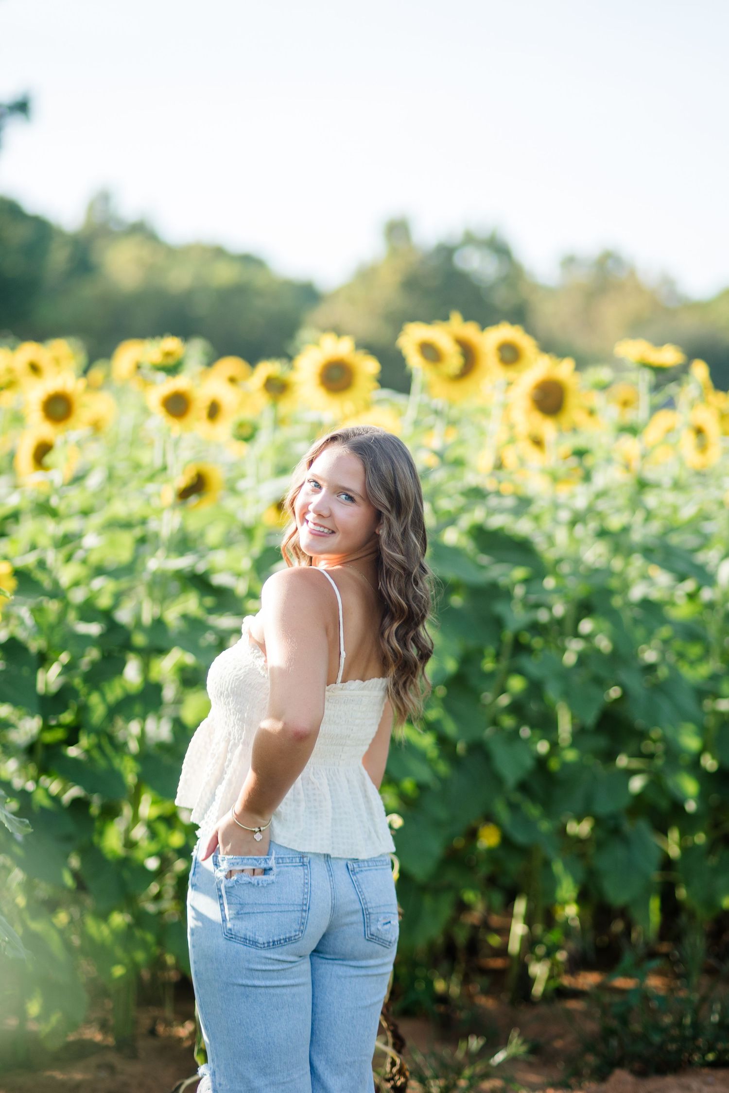 Young woman in white top and jeans smiling over shoulder in vibrant sunflower field on a sunny day.
