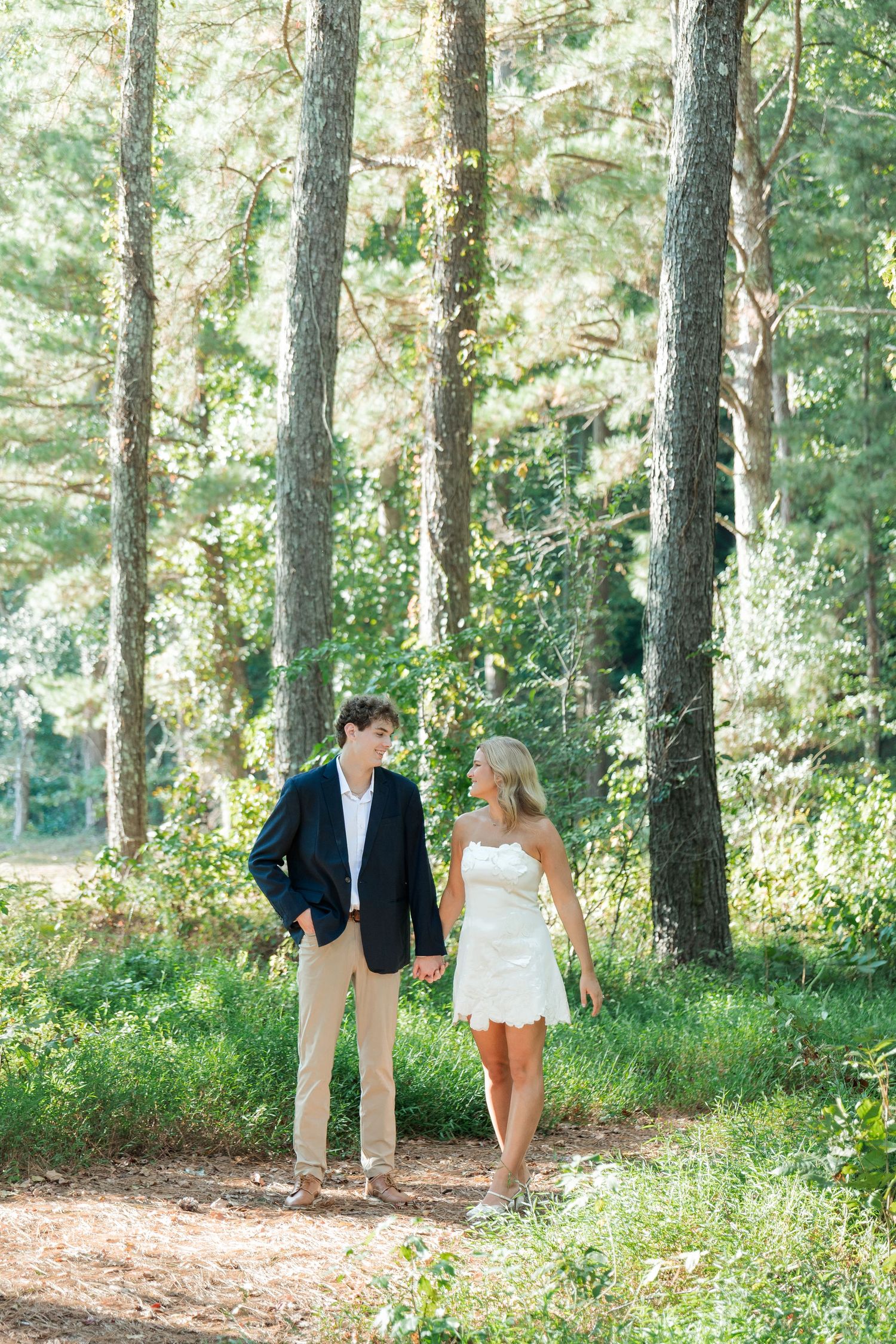Bride and groom strolling hand in hand on forest path surrounded by tall pine trees in sunlight.